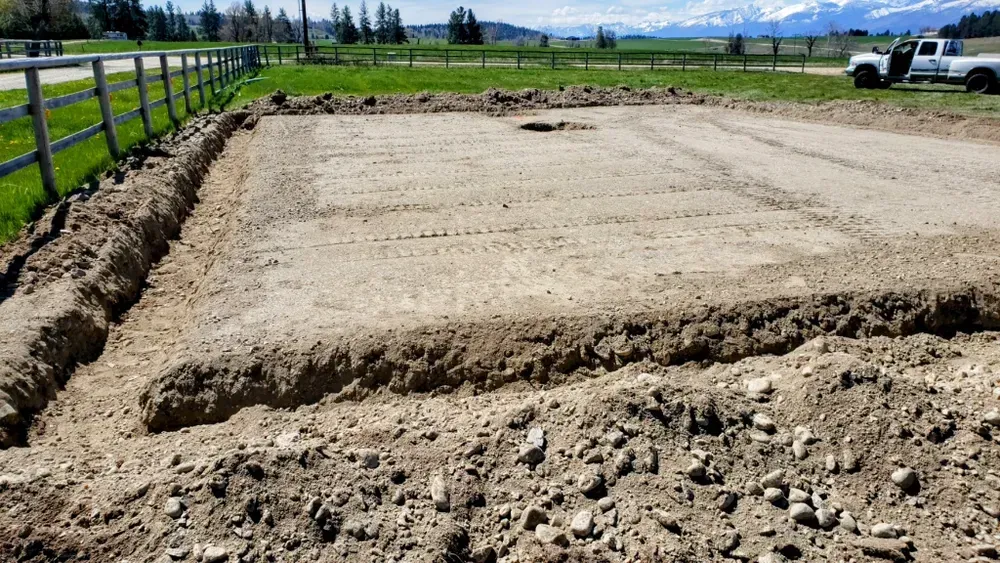 A rectangular foundation dug in dirt; a white fence, grass, and mountains in the background.