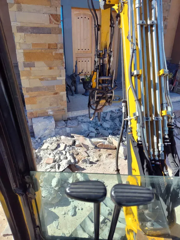 Inside excavator cab, jackhammer breaking concrete at doorway. Yellow equipment, light brick building.