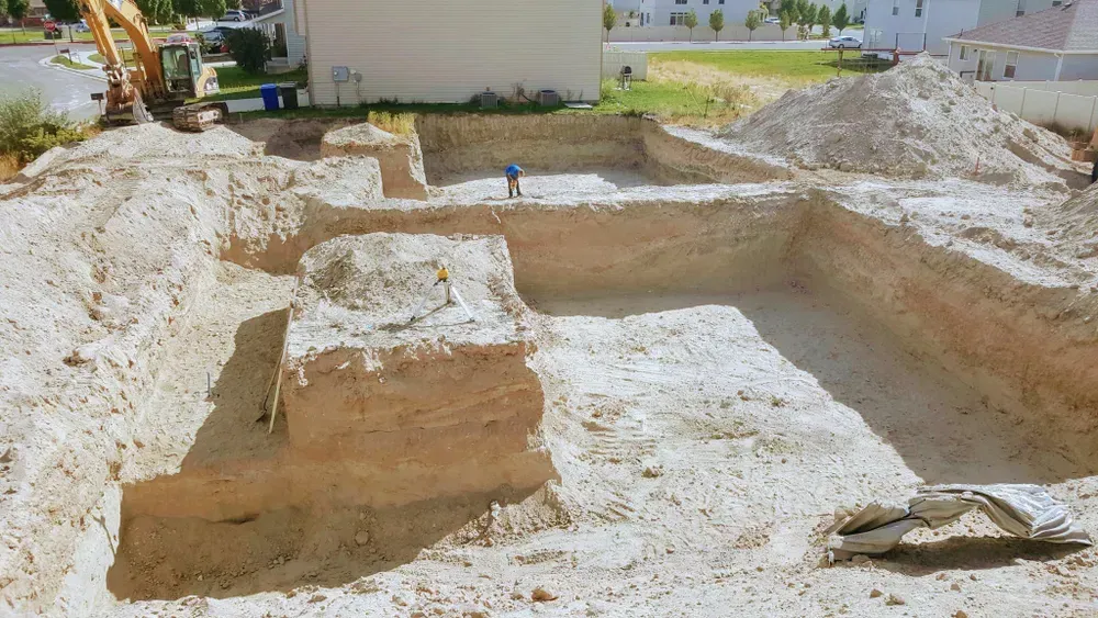 Excavation site with exposed soil walls. Backhoe, pile of dirt, and a person standing in the foundation.