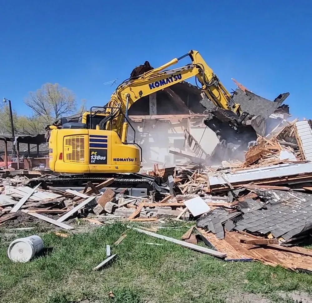 Yellow excavator demolishing a house on a sunny day; debris and a white bucket visible.
