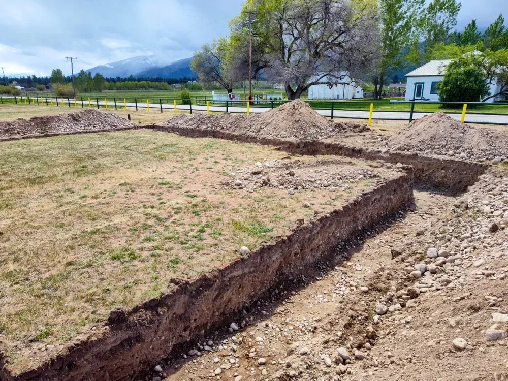 Excavated foundation trenches in a grassy field, with dirt piles and yellow markers. Mountains in the background.