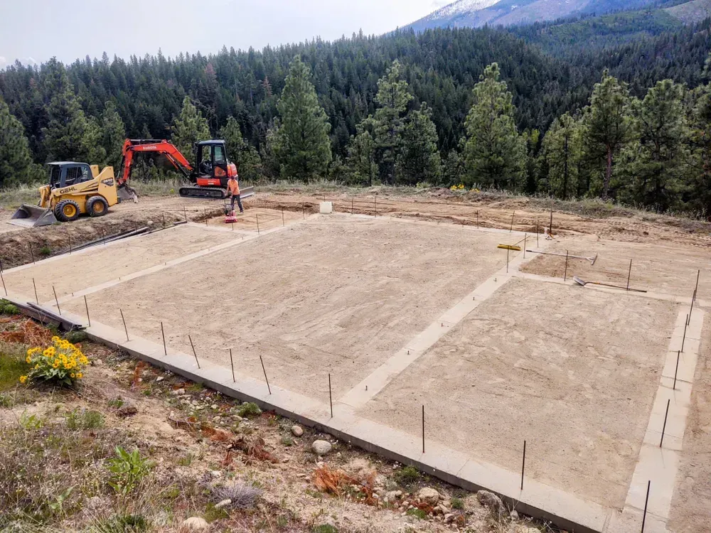 Construction site with an excavator, a compact track loader, and a concrete foundation. Forest and mountain backdrop.