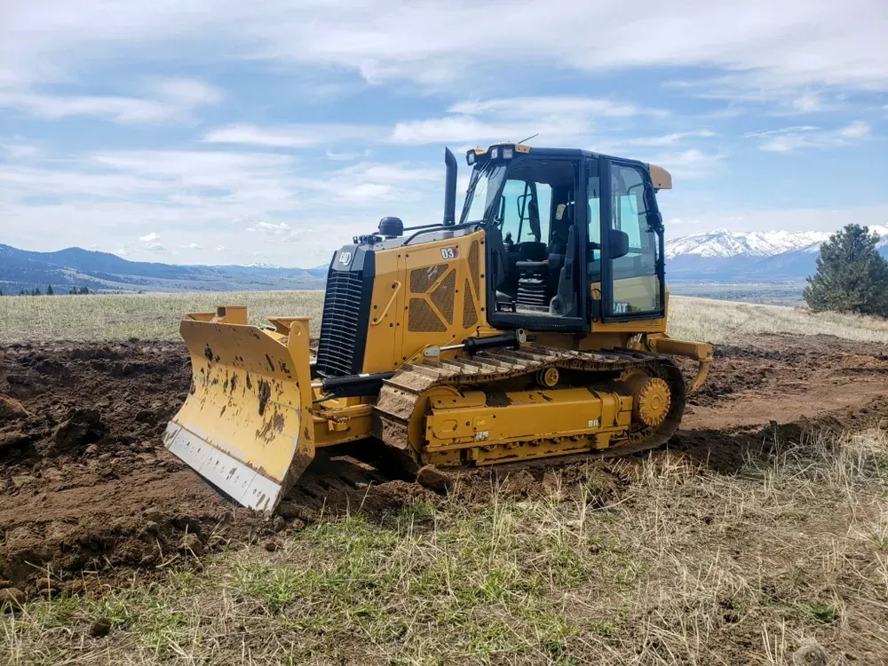 Yellow bulldozer plowing soil in a field, mountains and blue sky in the background.