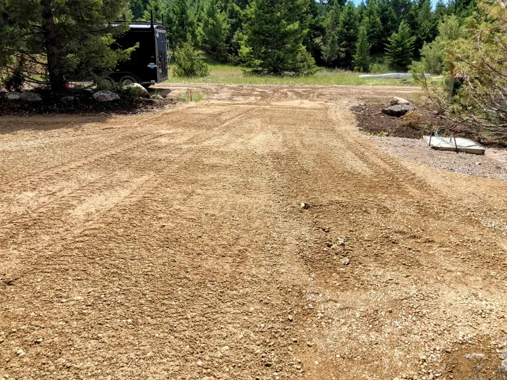 Gravel driveway with tire tracks, a parked trailer, and trees in the background.