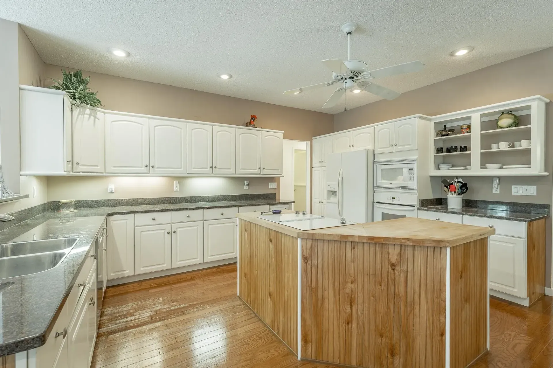 A kitchen with white cabinets , granite counter tops , a large island and a ceiling fan.