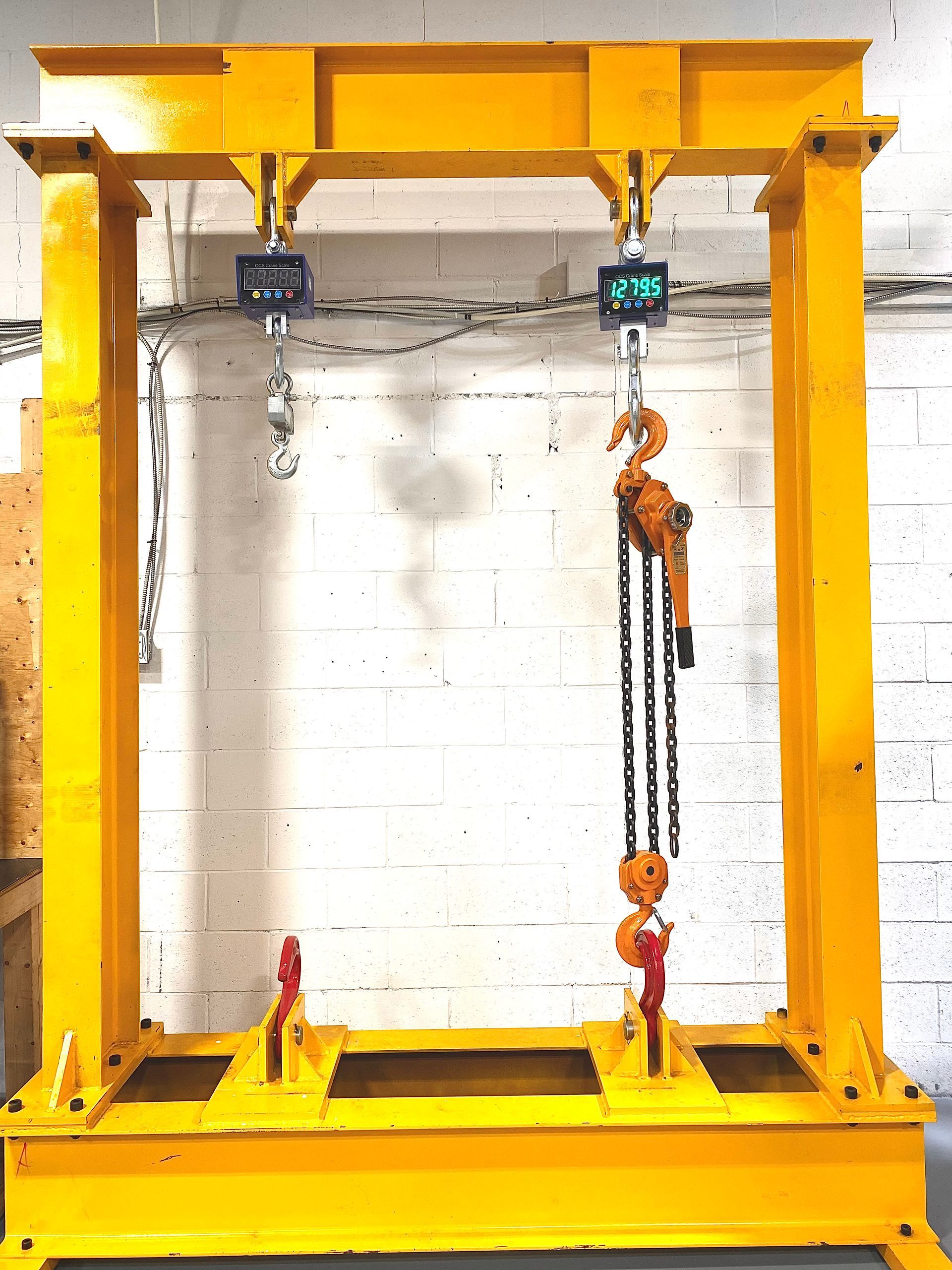 Cadre de levage en métal jaune avec deux balances suspendues et un palan à chaîne, contre un mur de briques blanches.