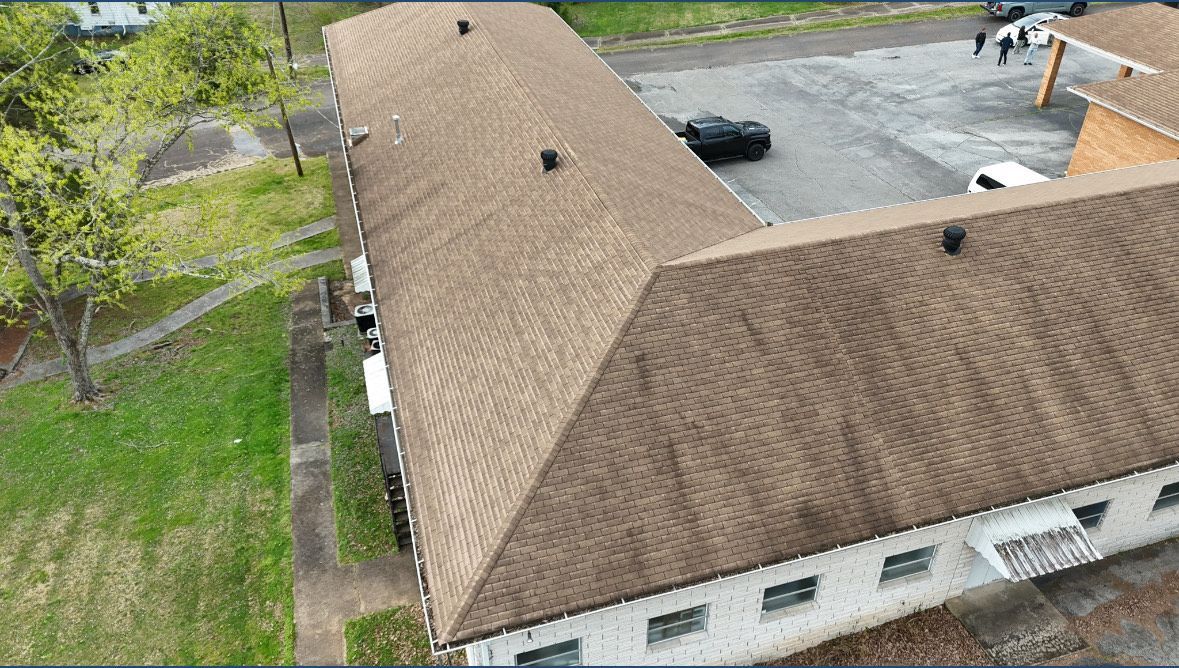 Aerial view of a light-colored building with a brown roof and surrounding green lawn and parking lot.