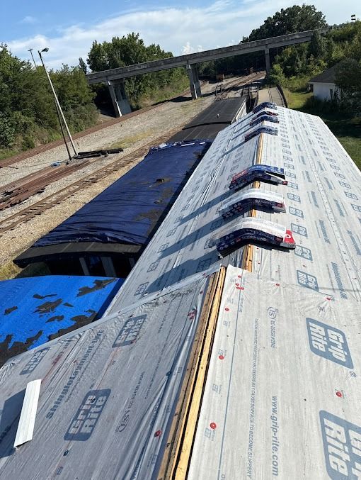 Roof of a building with partial new roof, and tarp over the older section. Rail tracks and overpass in the background.