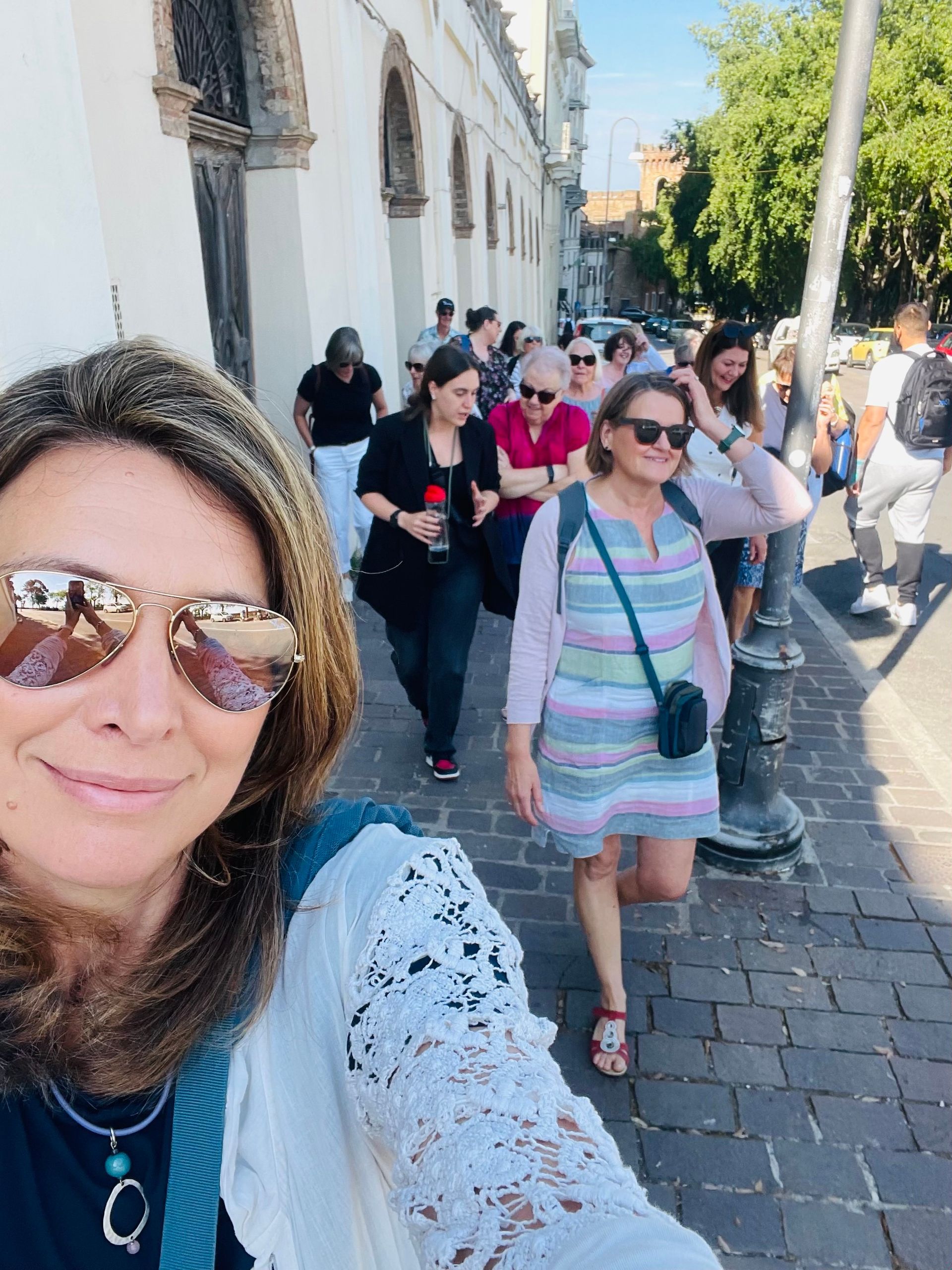 Woman taking selfie, group walking down a sunlit city street. People, building, and tree visible.