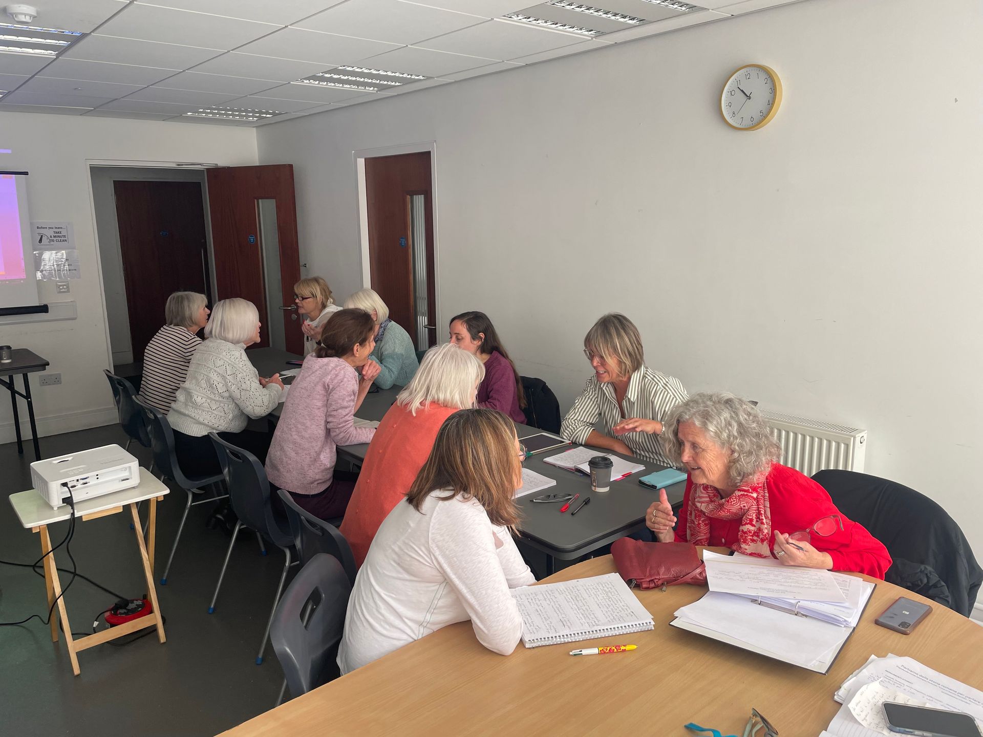 Group of women seated at tables in a room, engaged in conversation and writing.