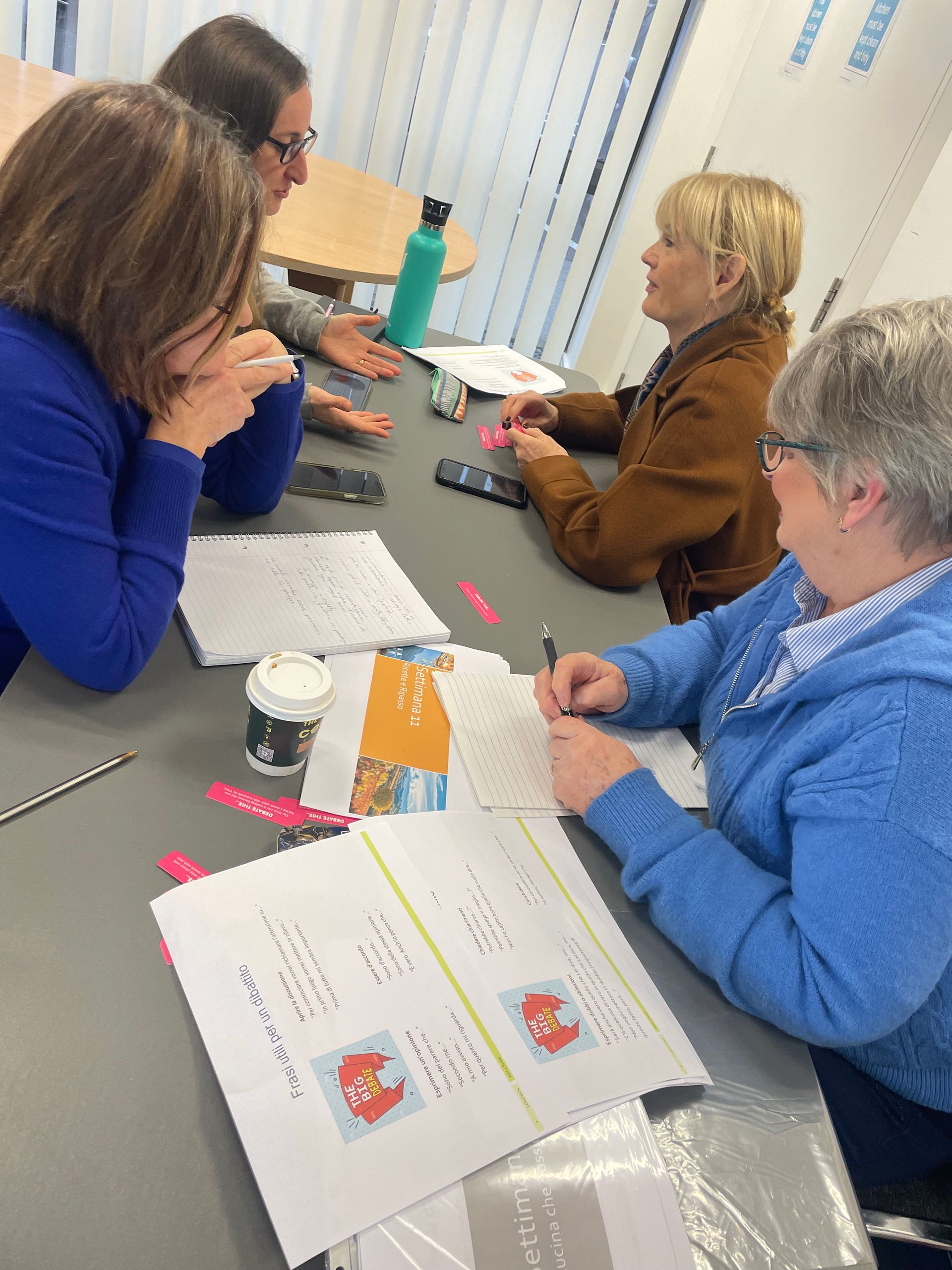 Four women at a table, working on papers, discussing. One is writing. Light setting.