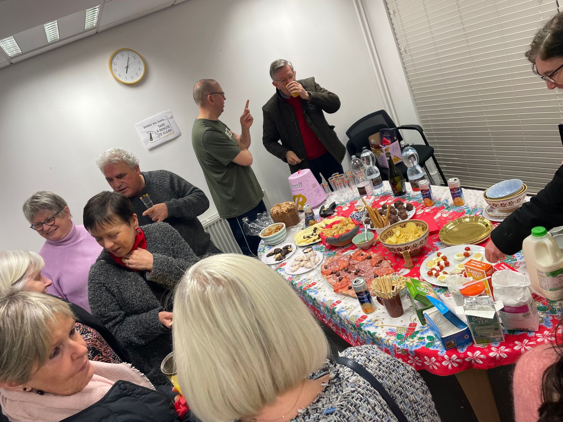 People gathered around a table laden with food and drinks. Some are chatting, smiling, and reaching for items.