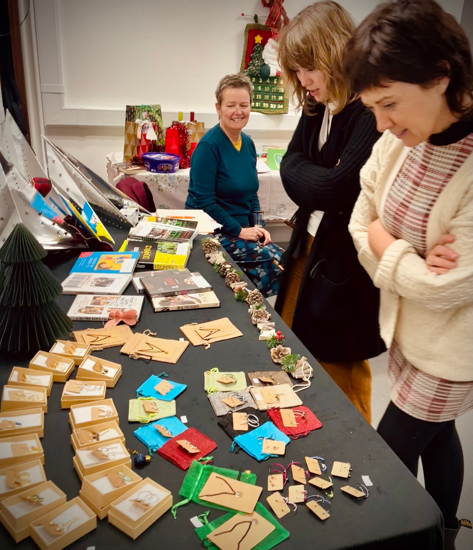 Women browsing crafts at a Christmas market. The vendor smiles, displaying jewelry and cards on a table.