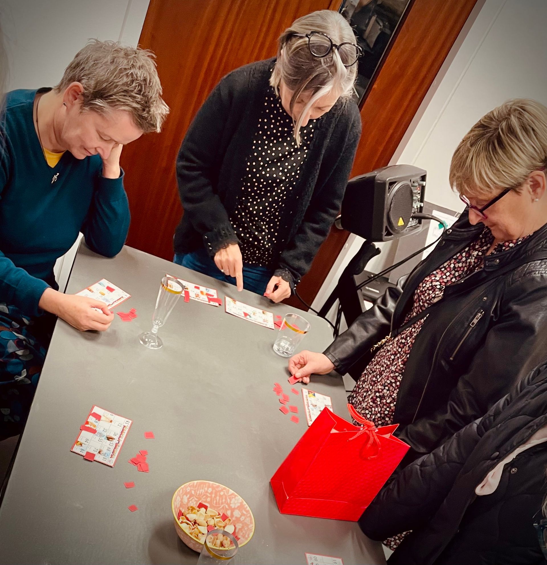 Three women play a game at a table. One points to cards; others examine theirs. A red gift bag sits nearby.