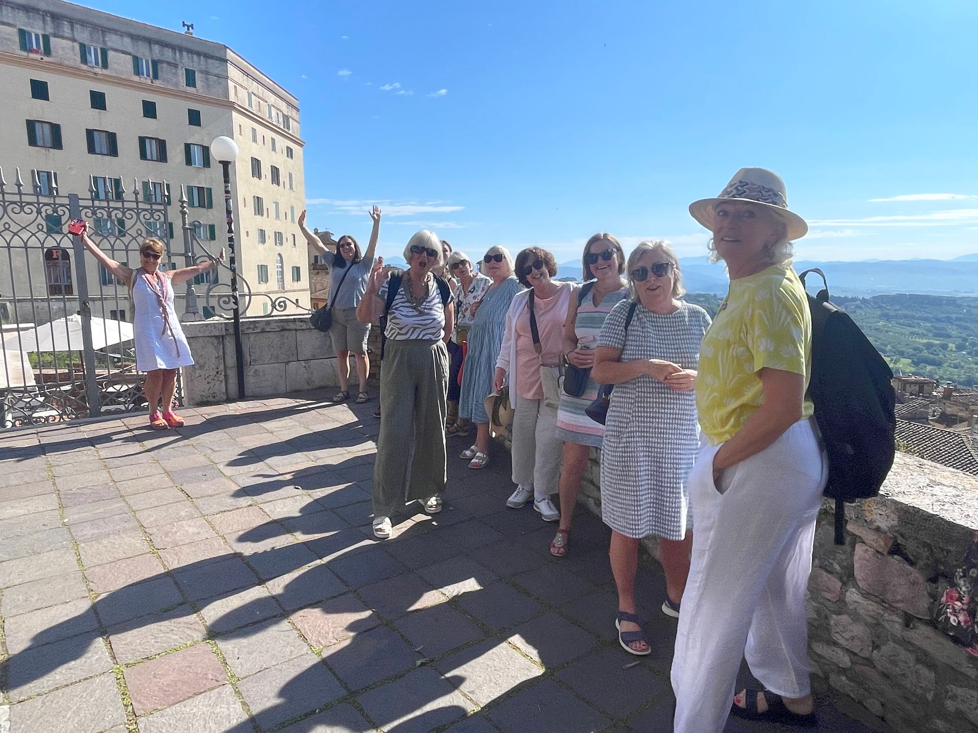 Group of tourists posing for a photo outside, sunny day. Some wearing hats. Tall building in background.