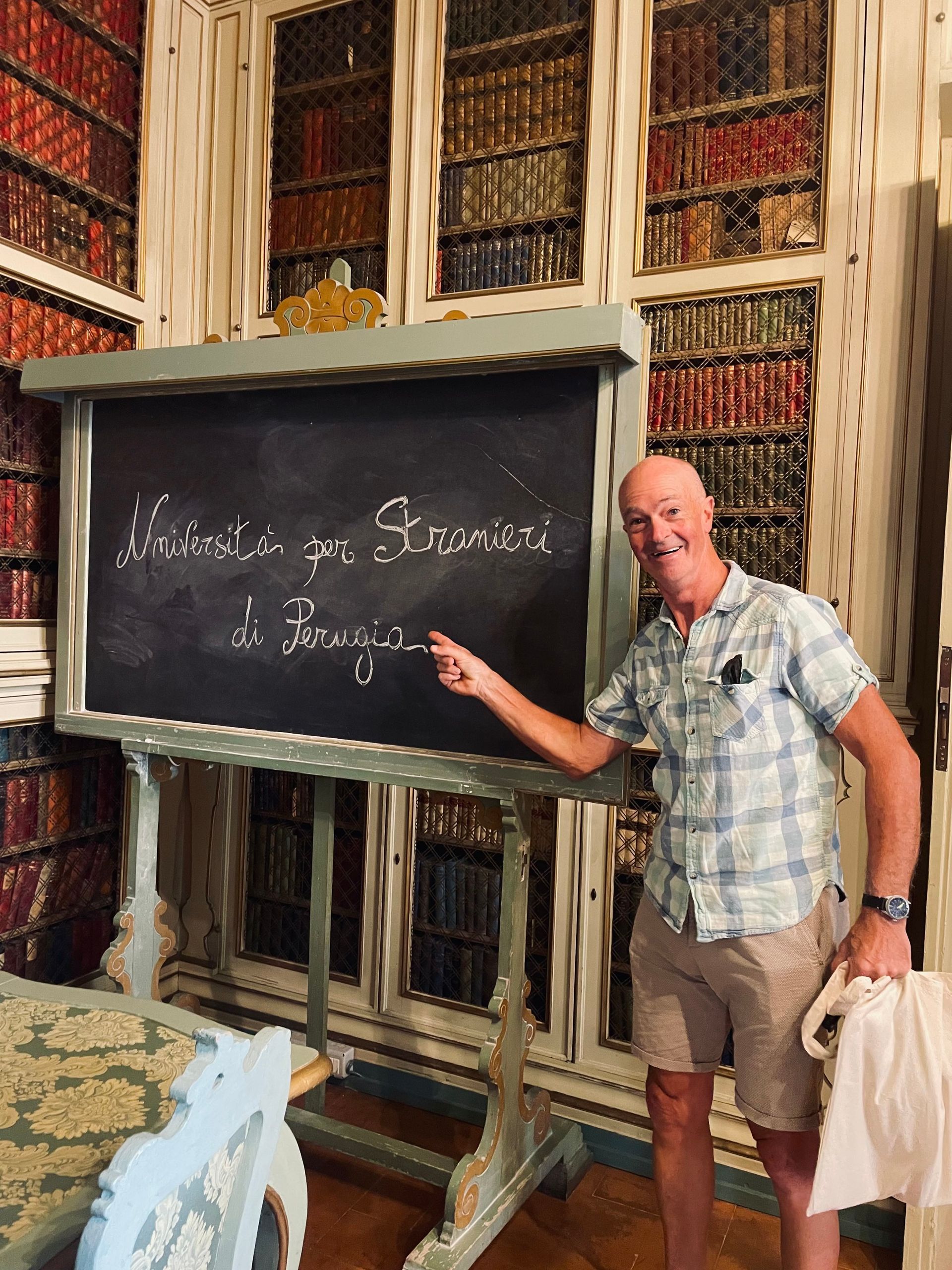 Man points to chalkboard with French inscription, standing in a library setting.