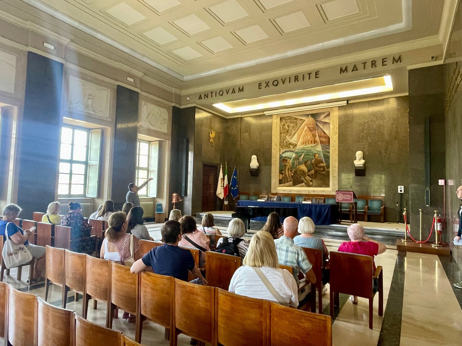 Group of people in a formal hall. A speaker stands near a table with flags. Large painting on the wall.