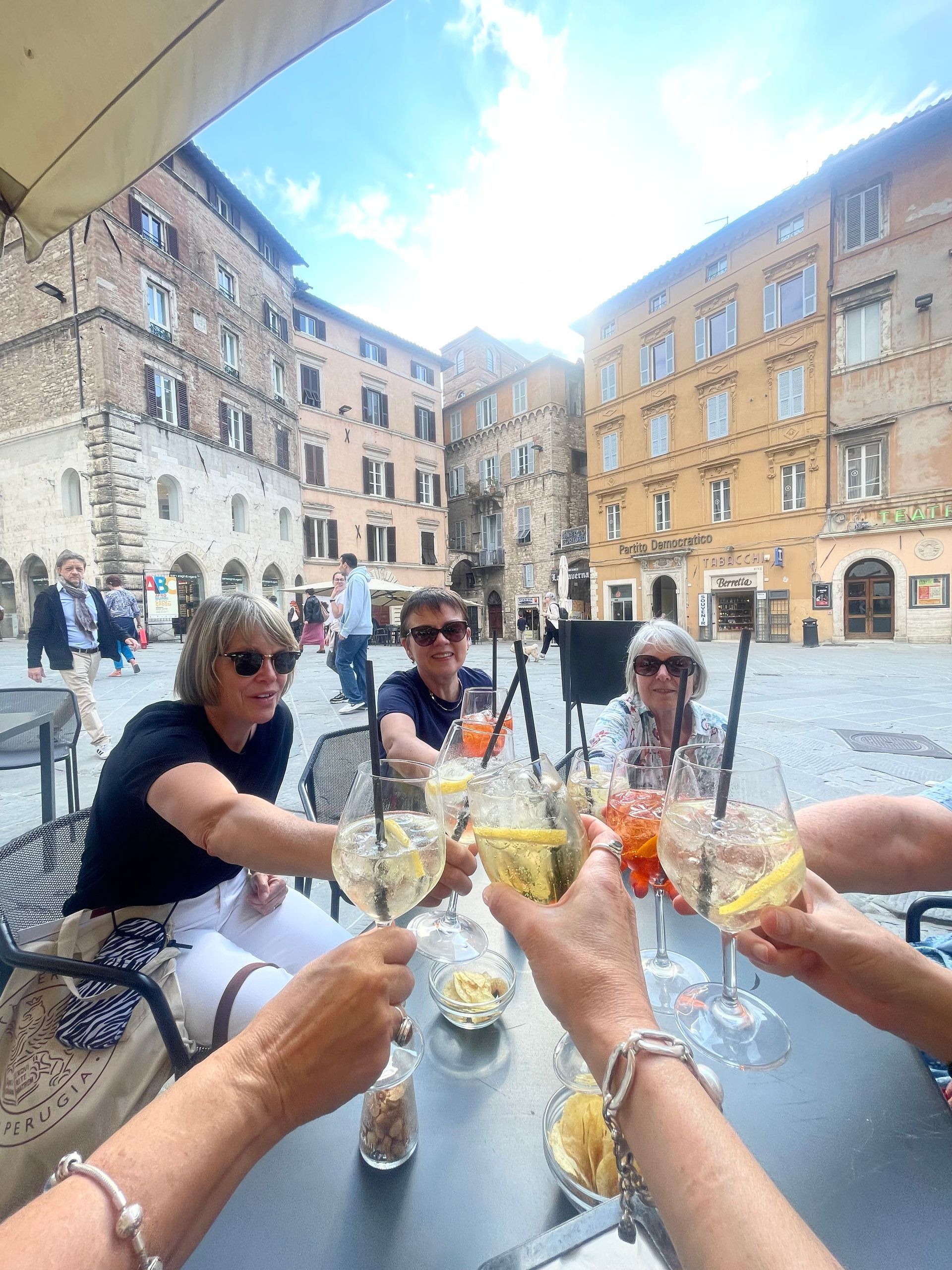 People toasting drinks outdoors in a European square. Buildings surround them. Sunny day.