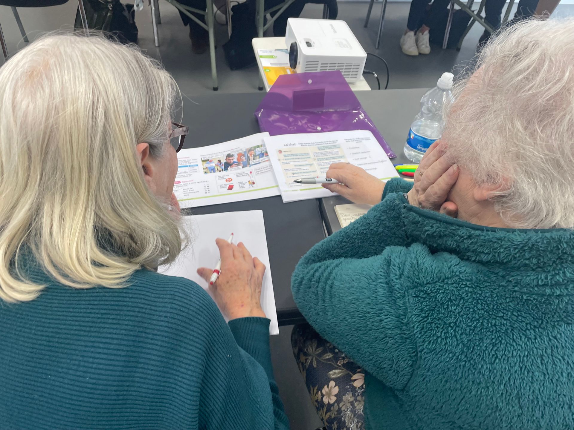 Two older women with white hair are seated at a table, one writing and the other covering their face.