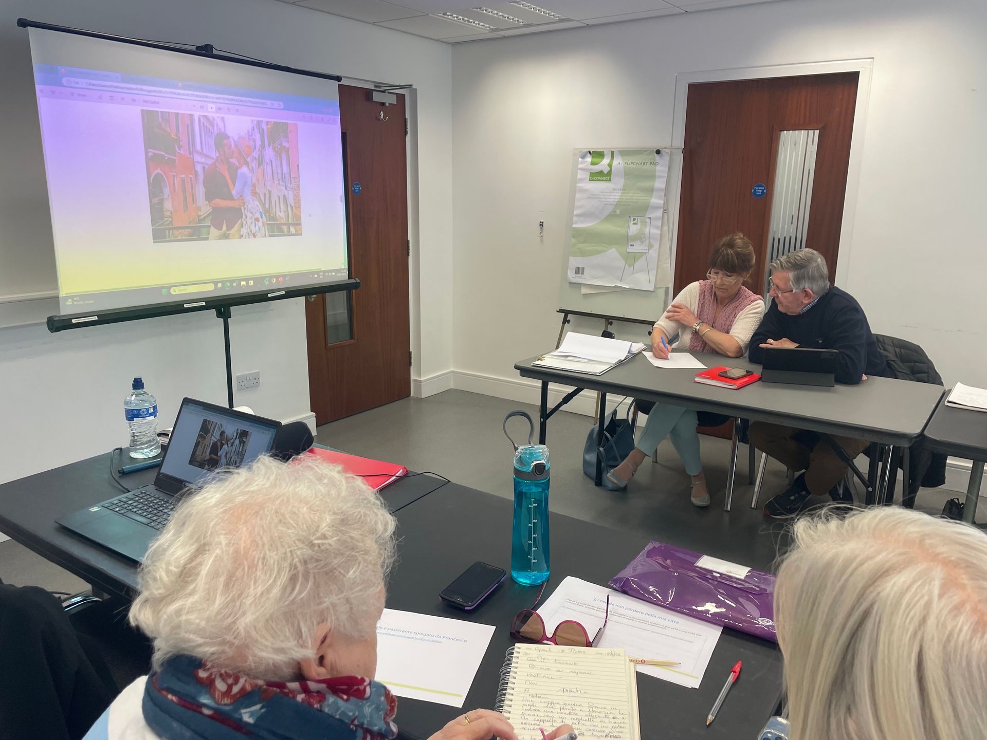 A meeting in a room with people looking at a presentation on a screen, taking notes.