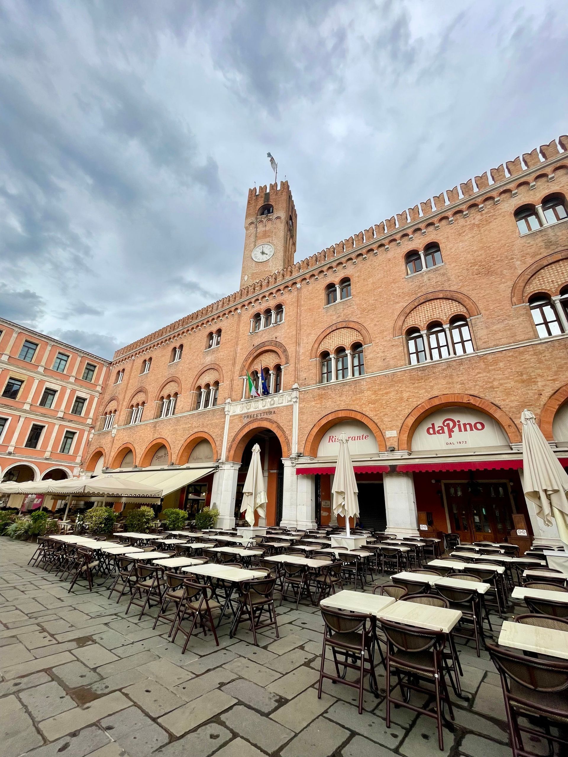 Exterior of a brick building with arched openings, a clock tower, and outdoor cafe seating. Overcast sky.