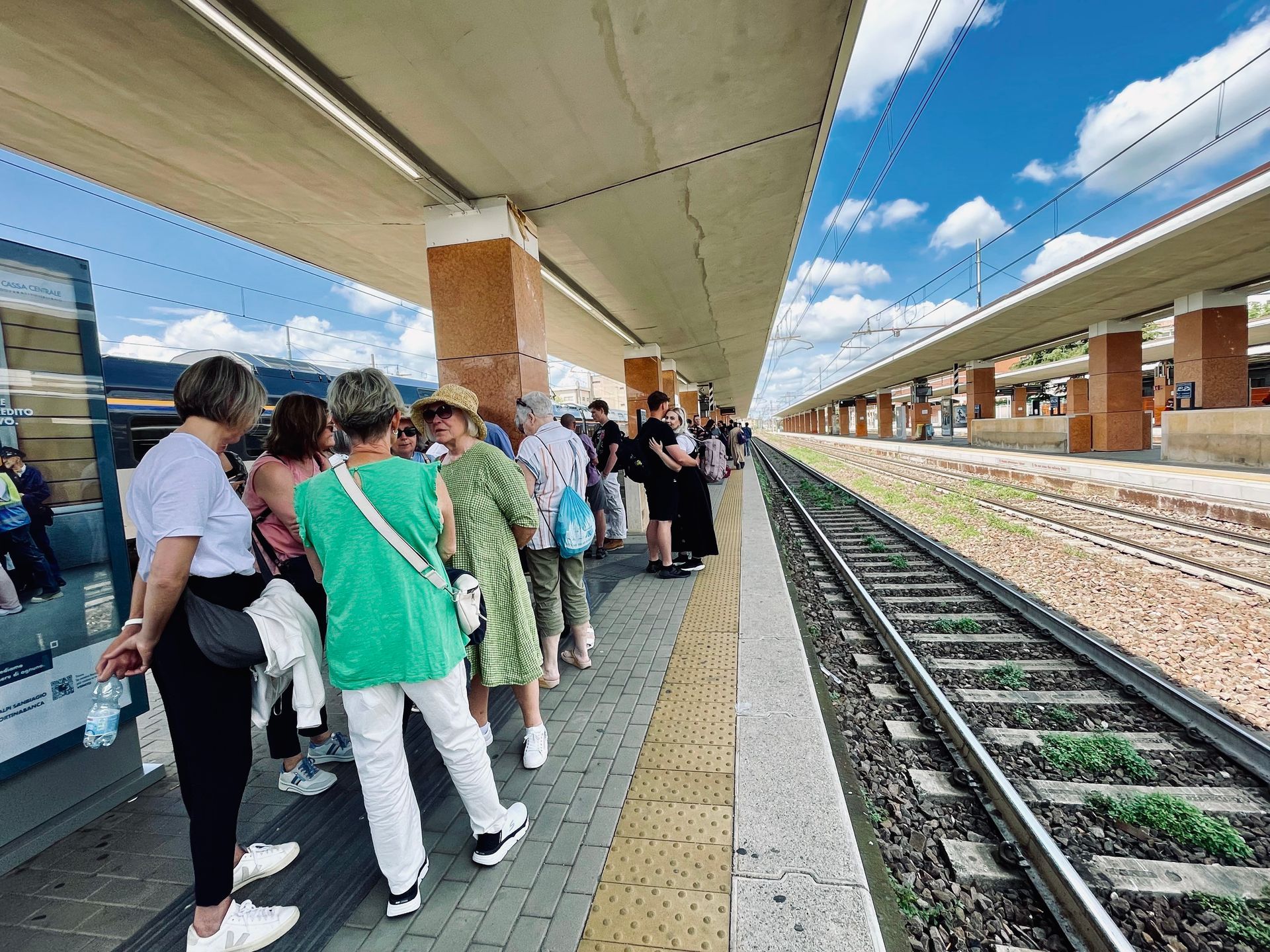 People wait on a train platform. Railroad tracks run beside the platform under a sunny sky.