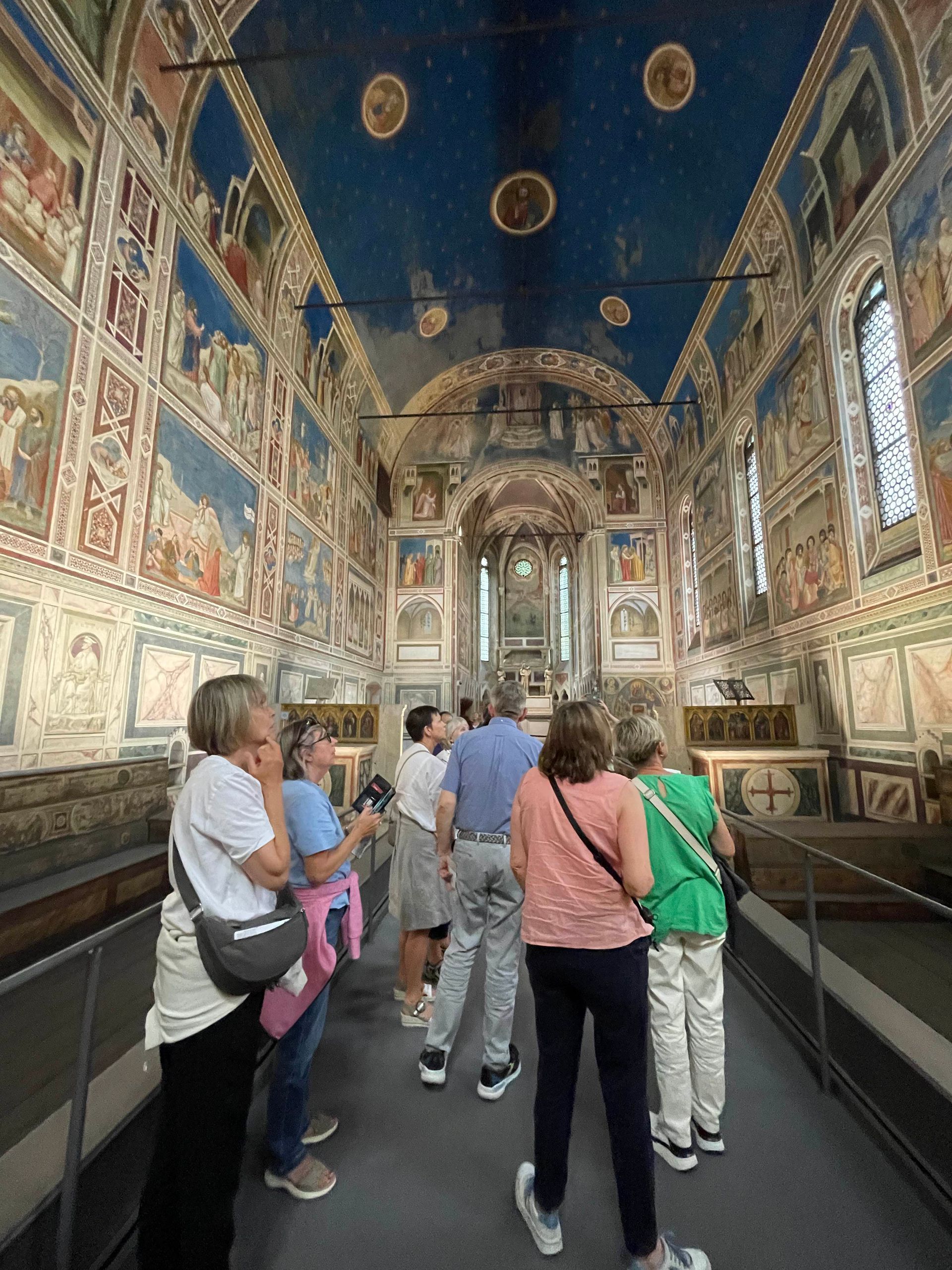People viewing the decorated interior of a chapel with frescoed walls and a blue ceiling.