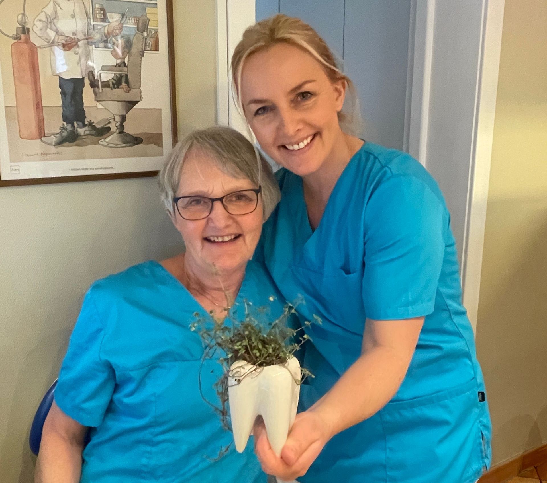 Tannhelsesekretærene Stine og Kari smiler mot kameraet mens de viser frem en potteplante formet som en tann