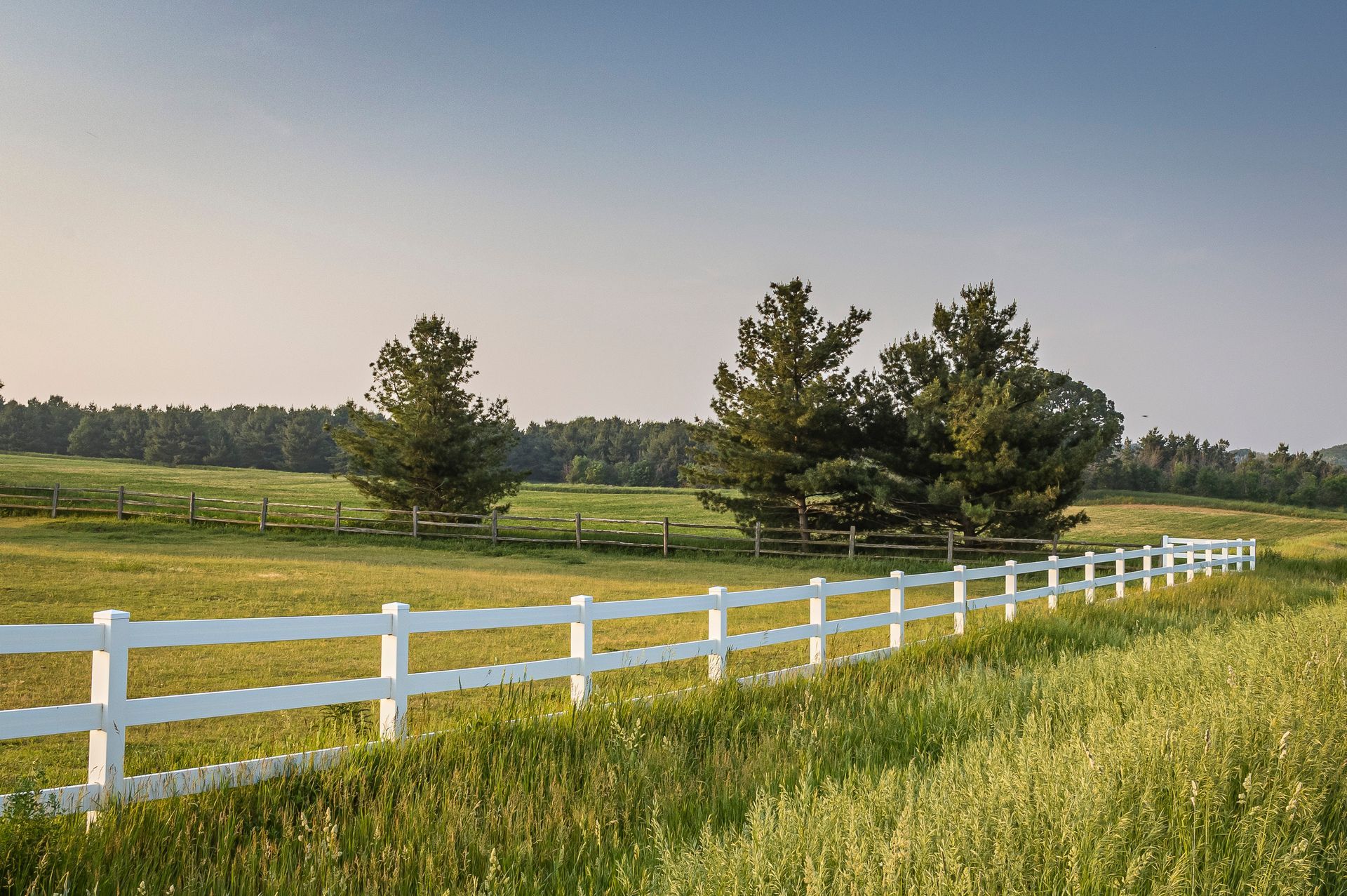 White fence beside a green meadow and pond under a clear blue sky