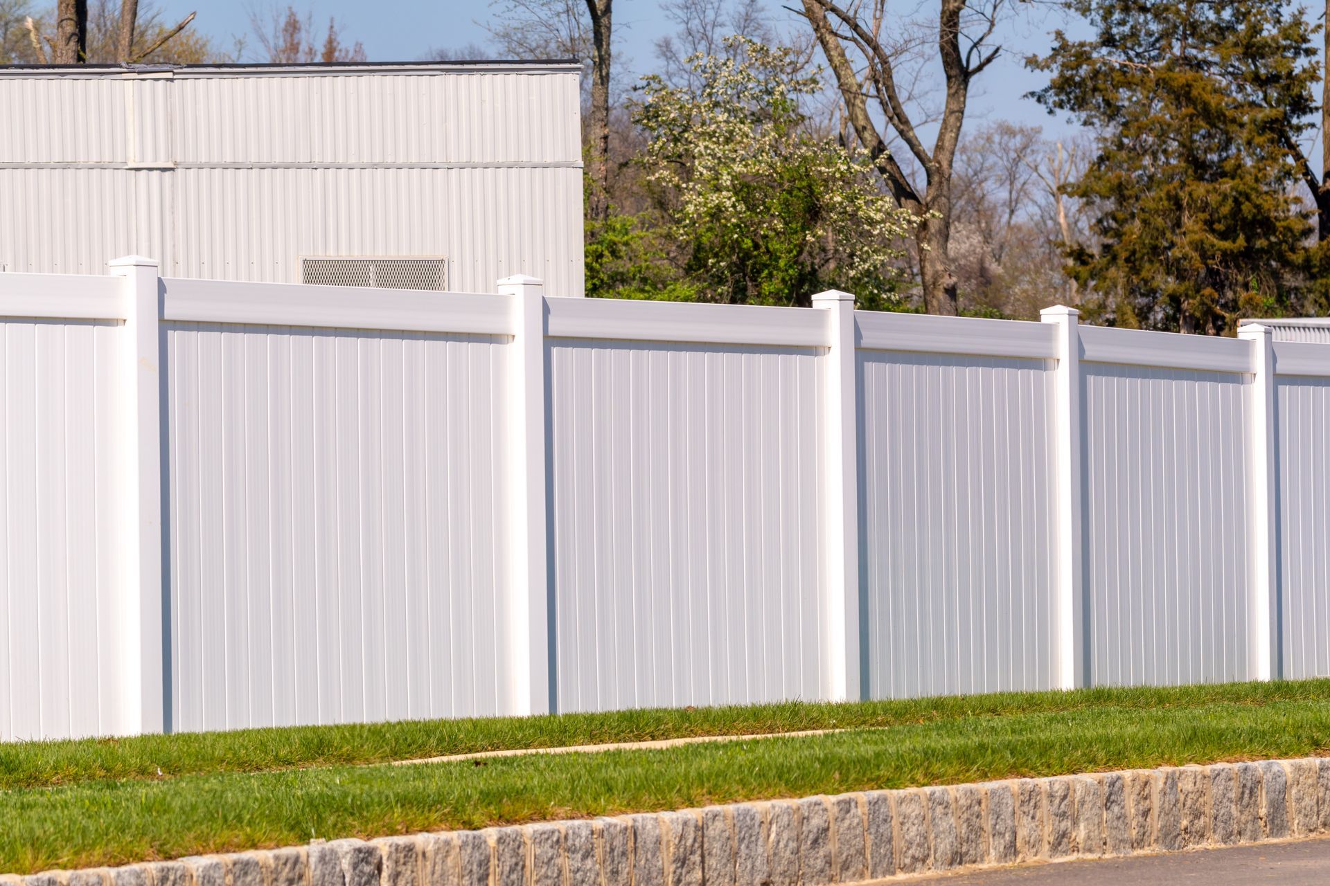 A white vinyl privacy fence lines a sidewalk with a green lawn and a stone border in front of a building.