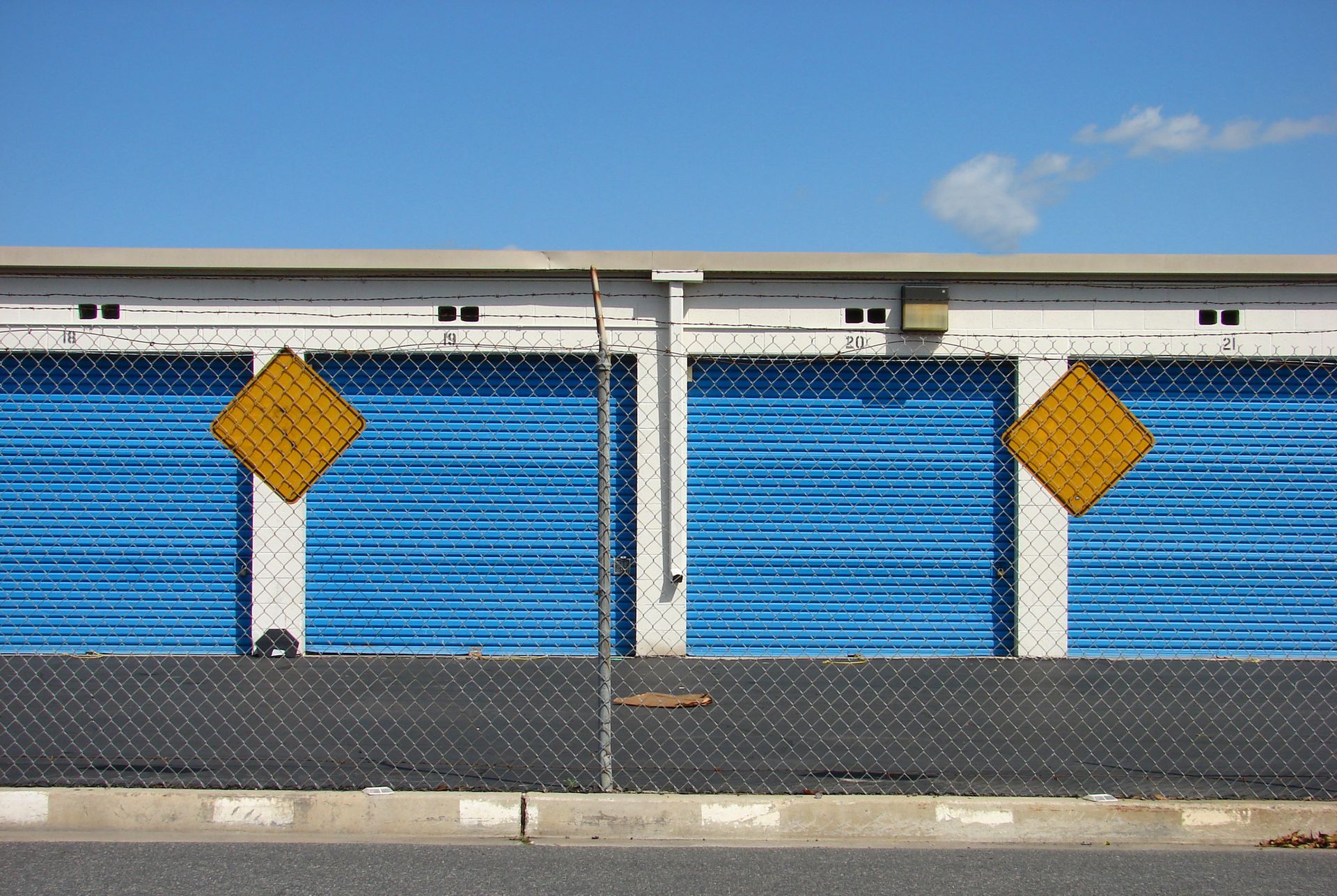 Storage facility with blue roll-up doors, chain-link fence, and two yellow warning signs against a clear blue sky. Storage facility with blue roll-up doors, chain-link fence, and two yellow warning signs against a clear blue sky.