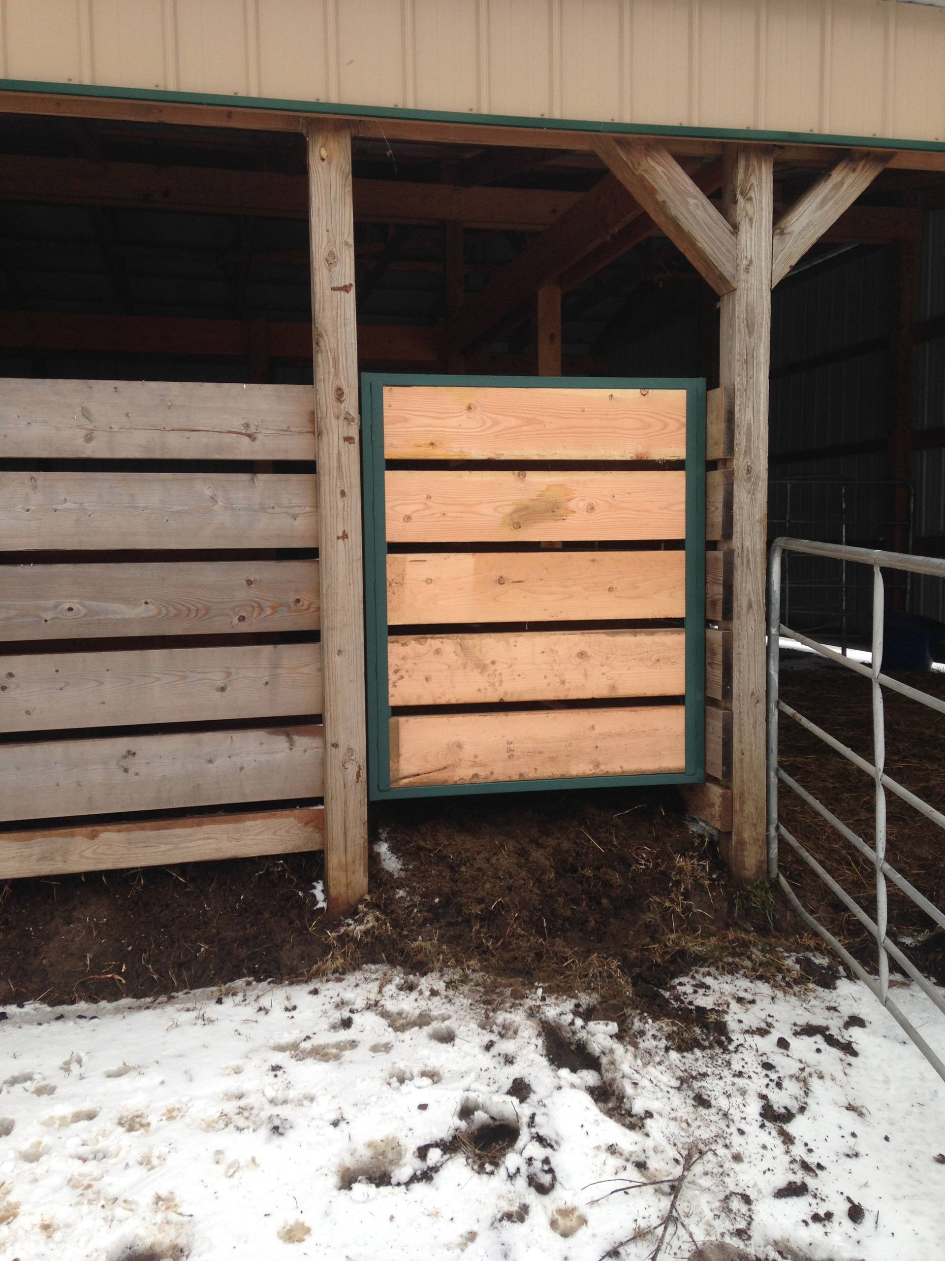 A wooden barn stall with a light tan gate, snow on the ground, and a metal fence on the right.