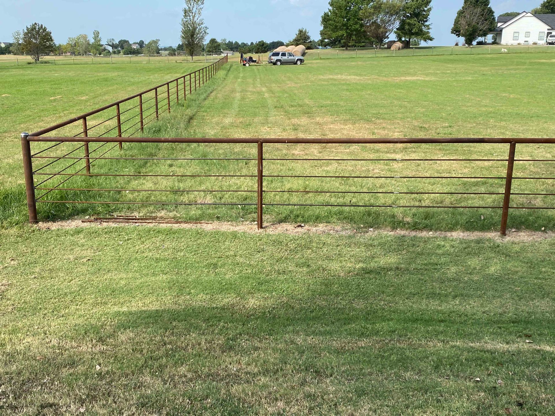 Rusty metal fence across a grassy field with trees and a distant house under a clear sky