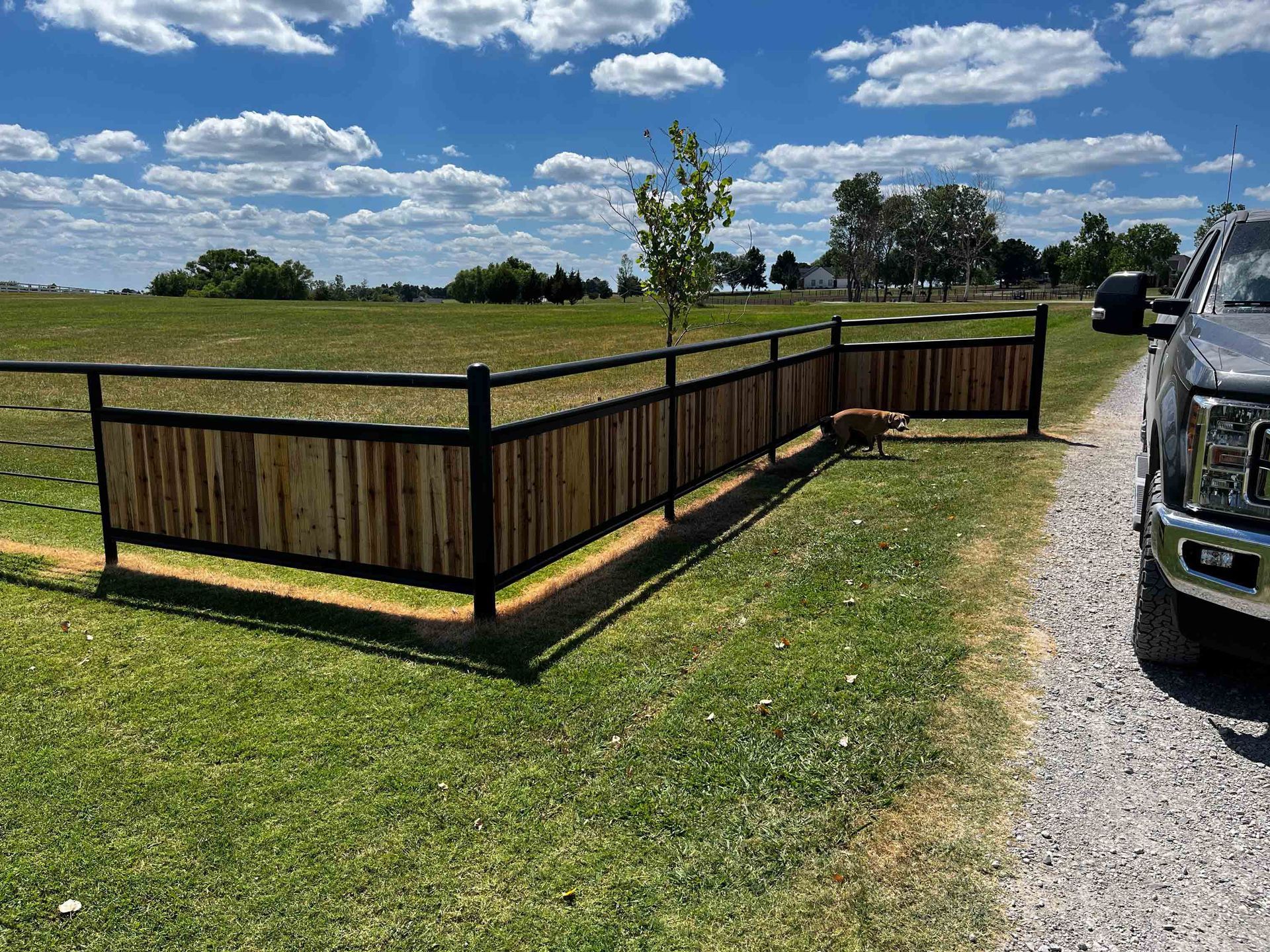 Wooden fenced enclosure in a grassy field beside a gravel road under a blue sky with clouds