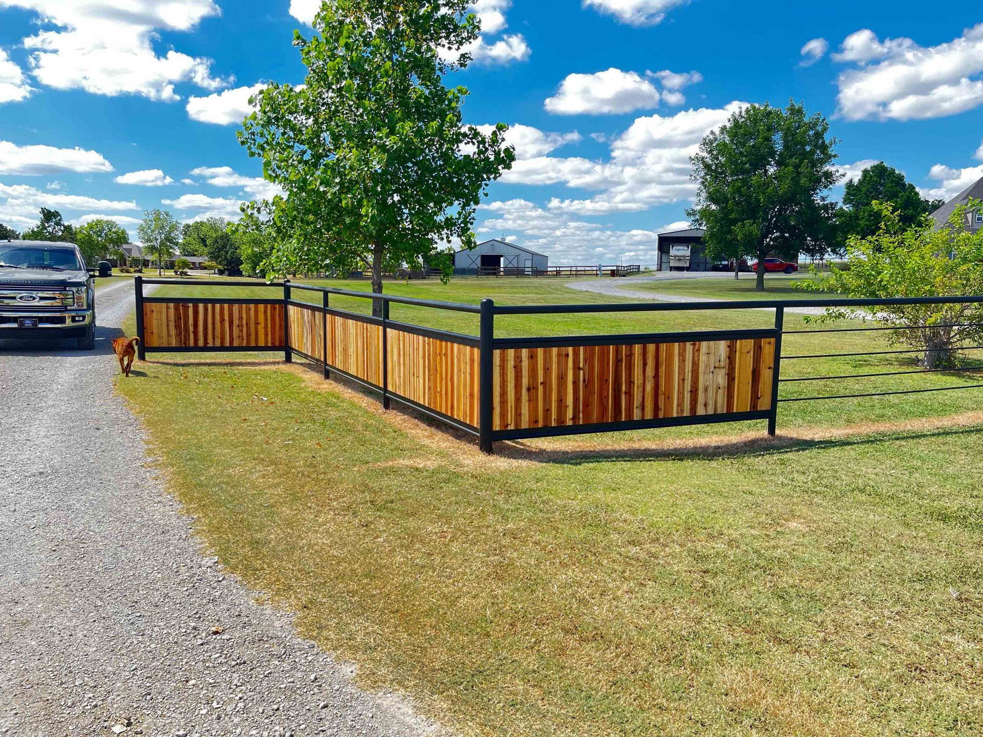 Gravel driveway beside a wooden fence around a grassy field, with a tree, truck, and blue sky with clouds.