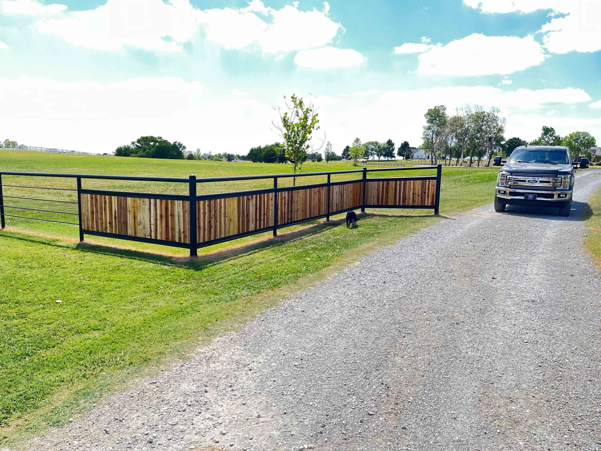 Gravel driveway beside a wooden fence and grassy field, with a parked black SUV under a bright blue sky.