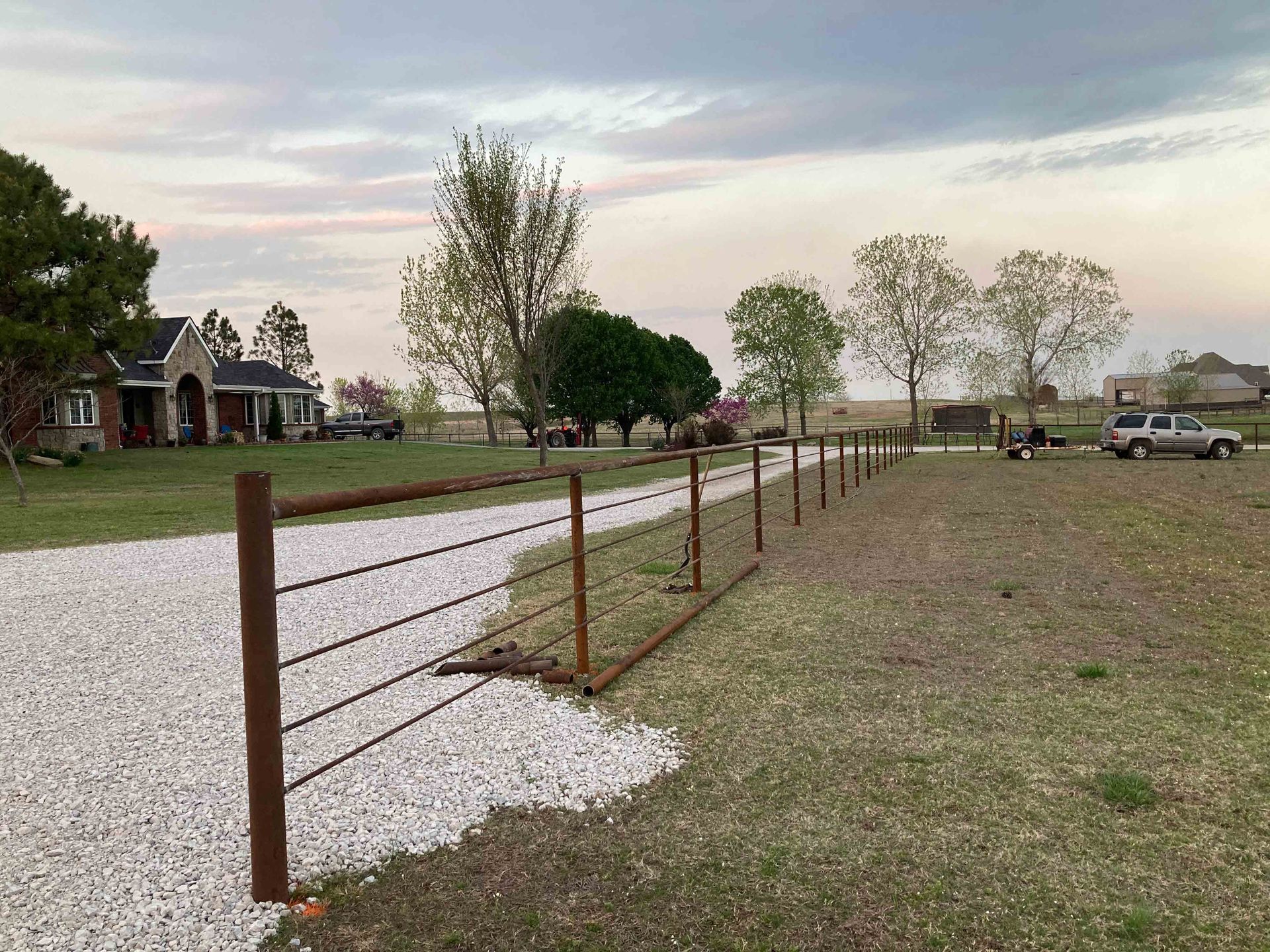 Rural gravel driveway beside a wire fence, with a house, trees, and parked pickup under a cloudy sky.
