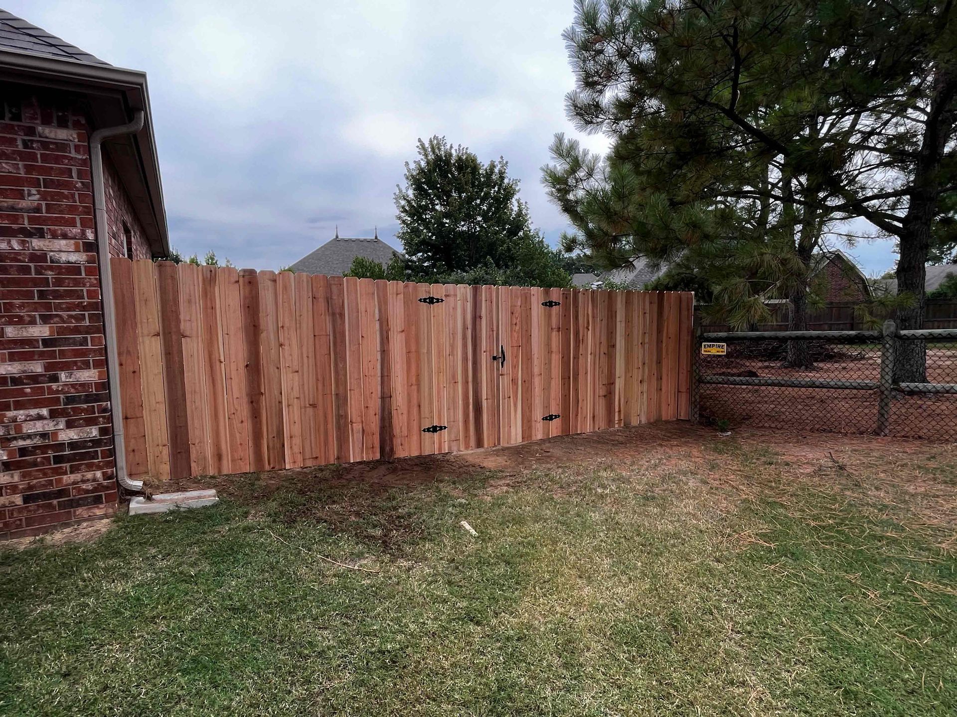 Wooden privacy fence along a grassy yard beside a brick house, with trees and a chain-link fence in back.