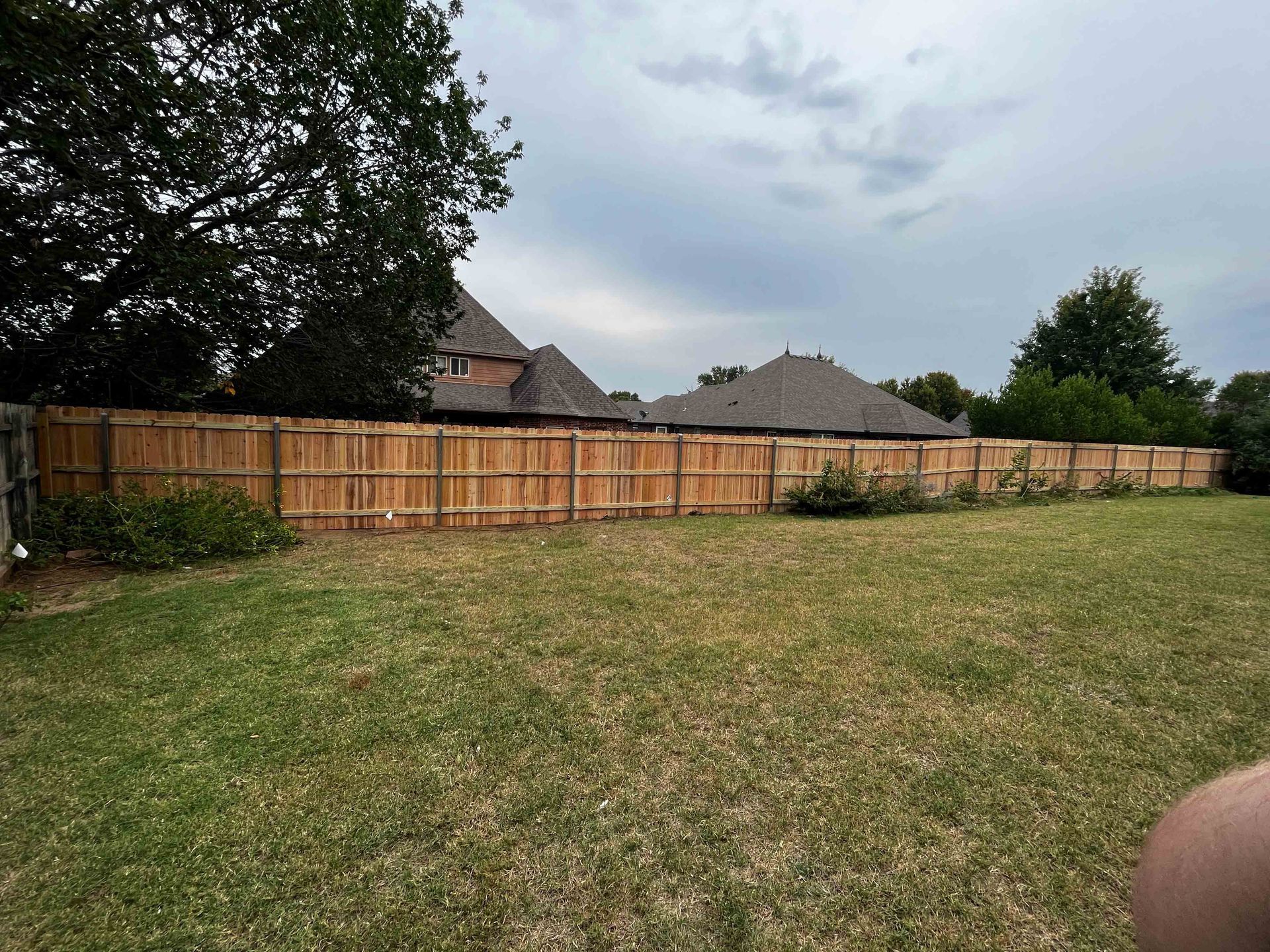 Wide grassy yard with a wooden fence, trees, and rooftops under a cloudy sky