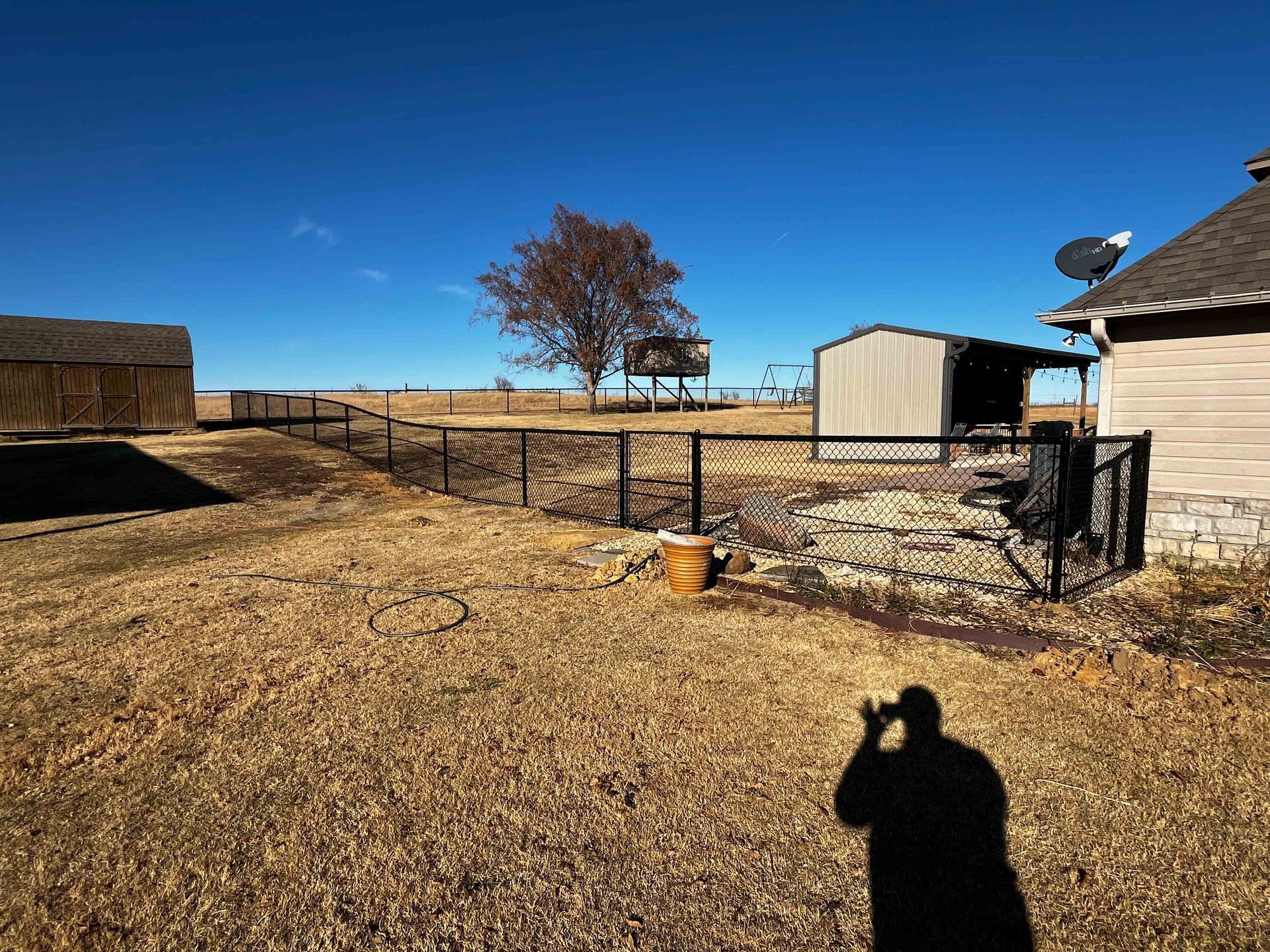 Rural yard with fence, shed, lone tree, and a person's shadow on dry ground under a bright blue sky