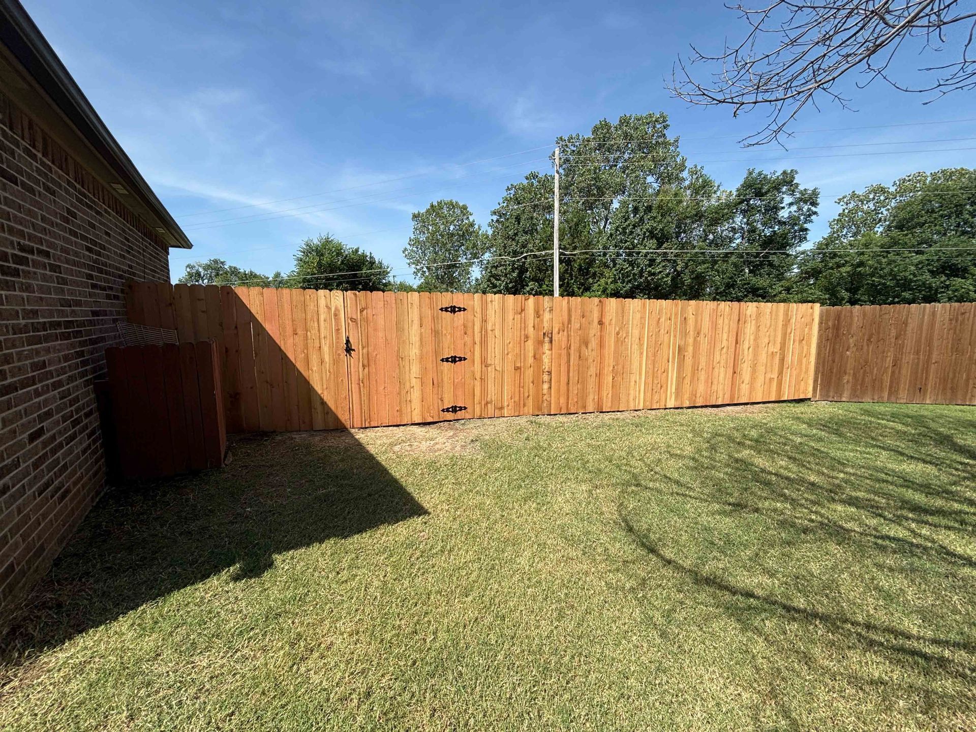 Backyard with wooden privacy fence, green lawn, brick house side, and blue sky