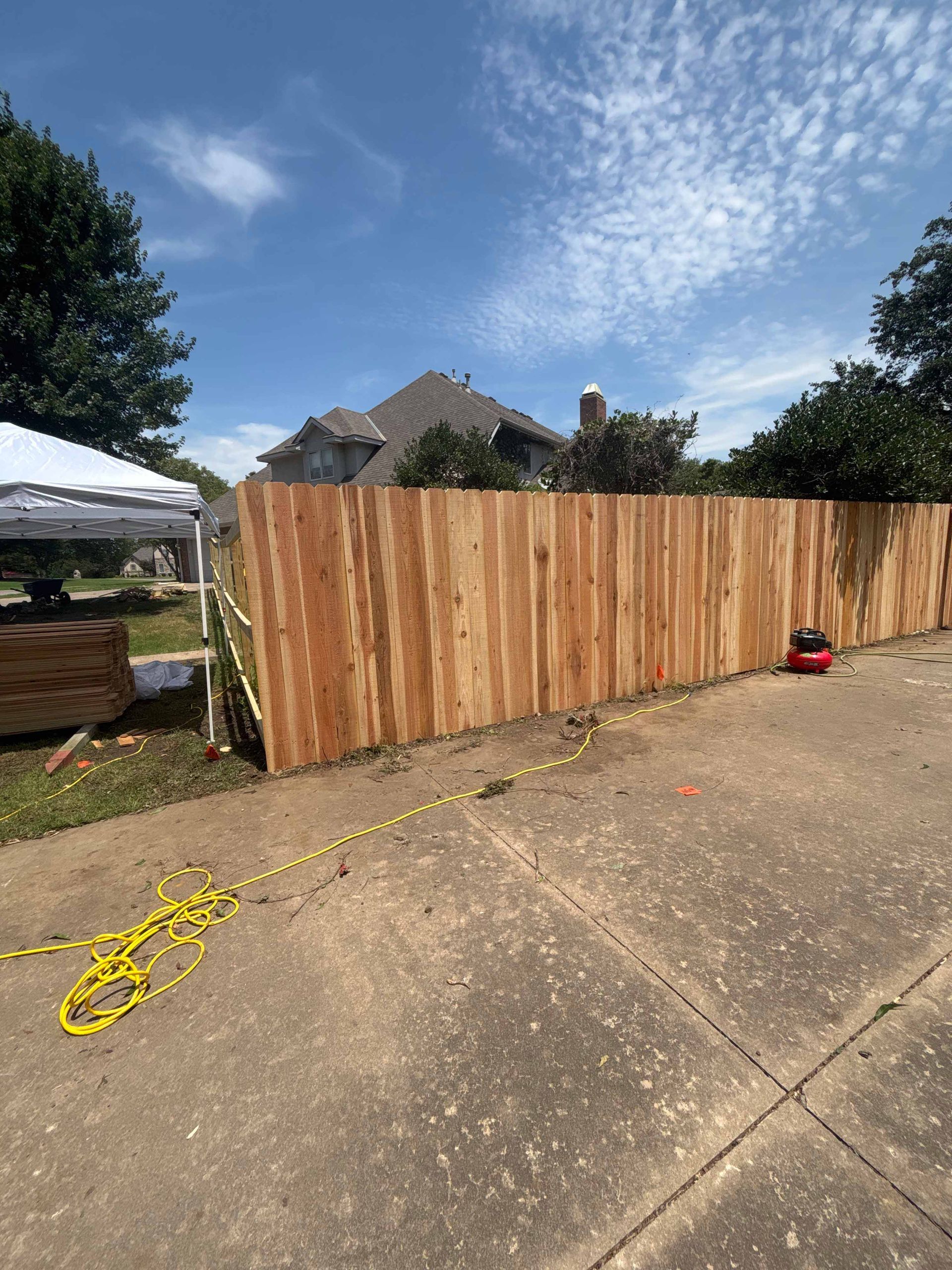 Wooden fence along a driveway under a blue sky, with a yellow cord and small red tool nearby.