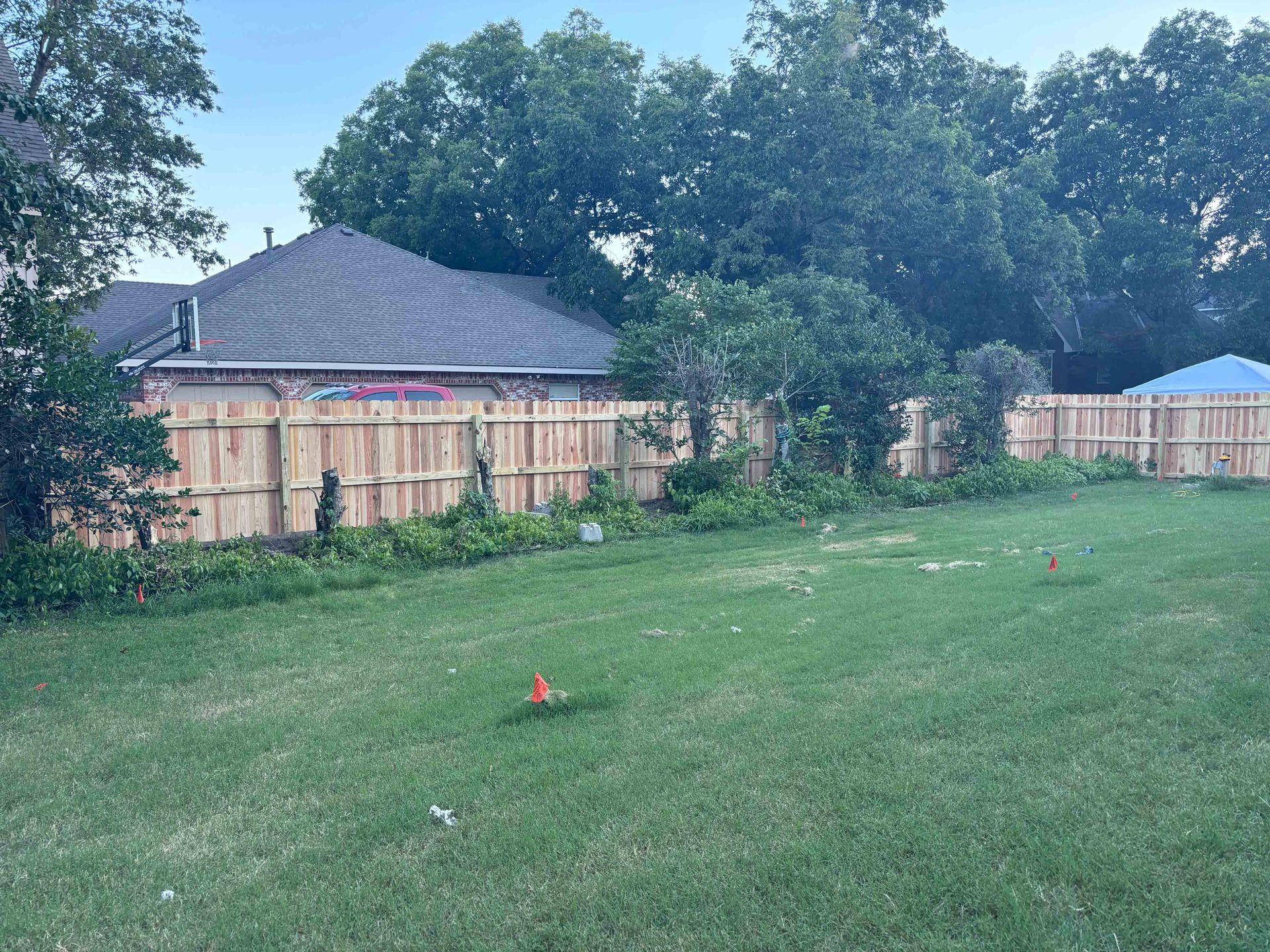 Grassy backyard with a wooden fence, trees, and houses under a cloudy sky