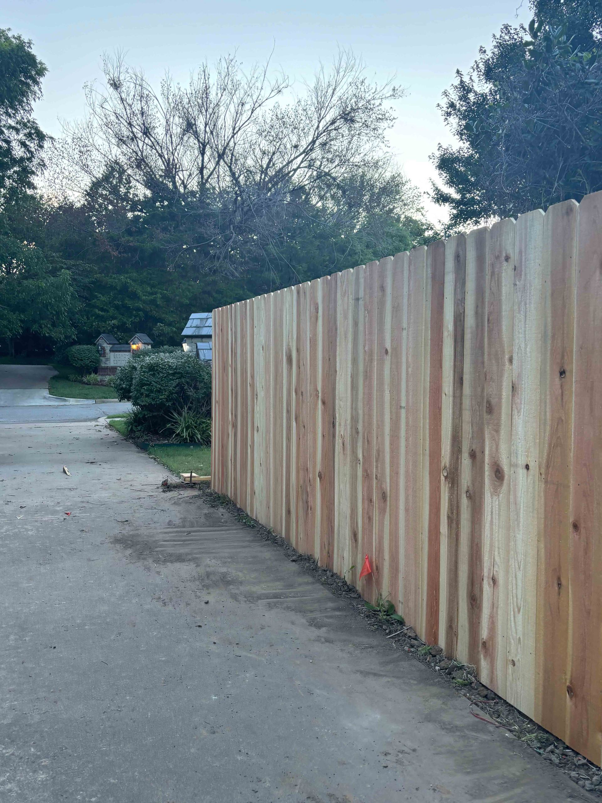 Wooden privacy fence along a paved path with trees and shrubs in the background.