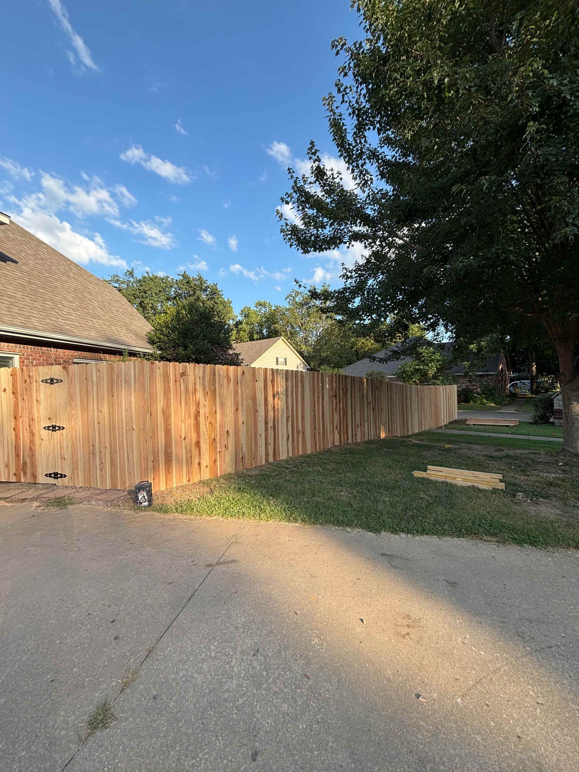 Wooden fence beside a gravel driveway under blue sky, with trees and a house roof nearby