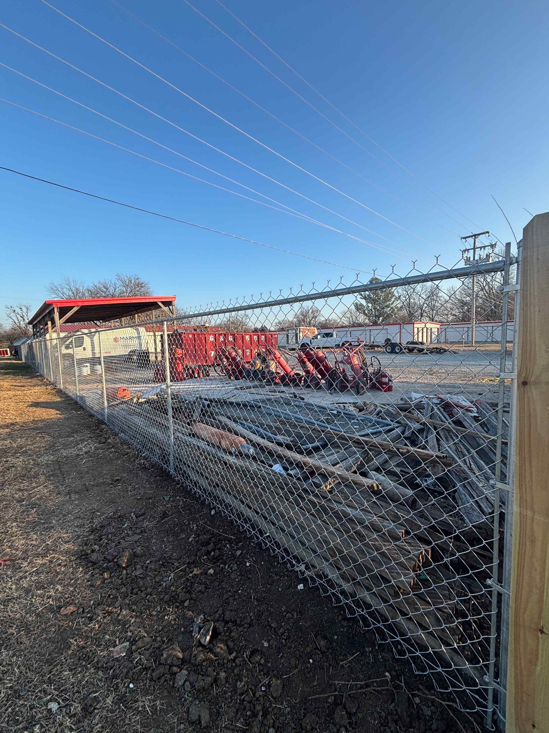 Chain-link fence in front of red tractors and farm equipment under a clear blue sky.