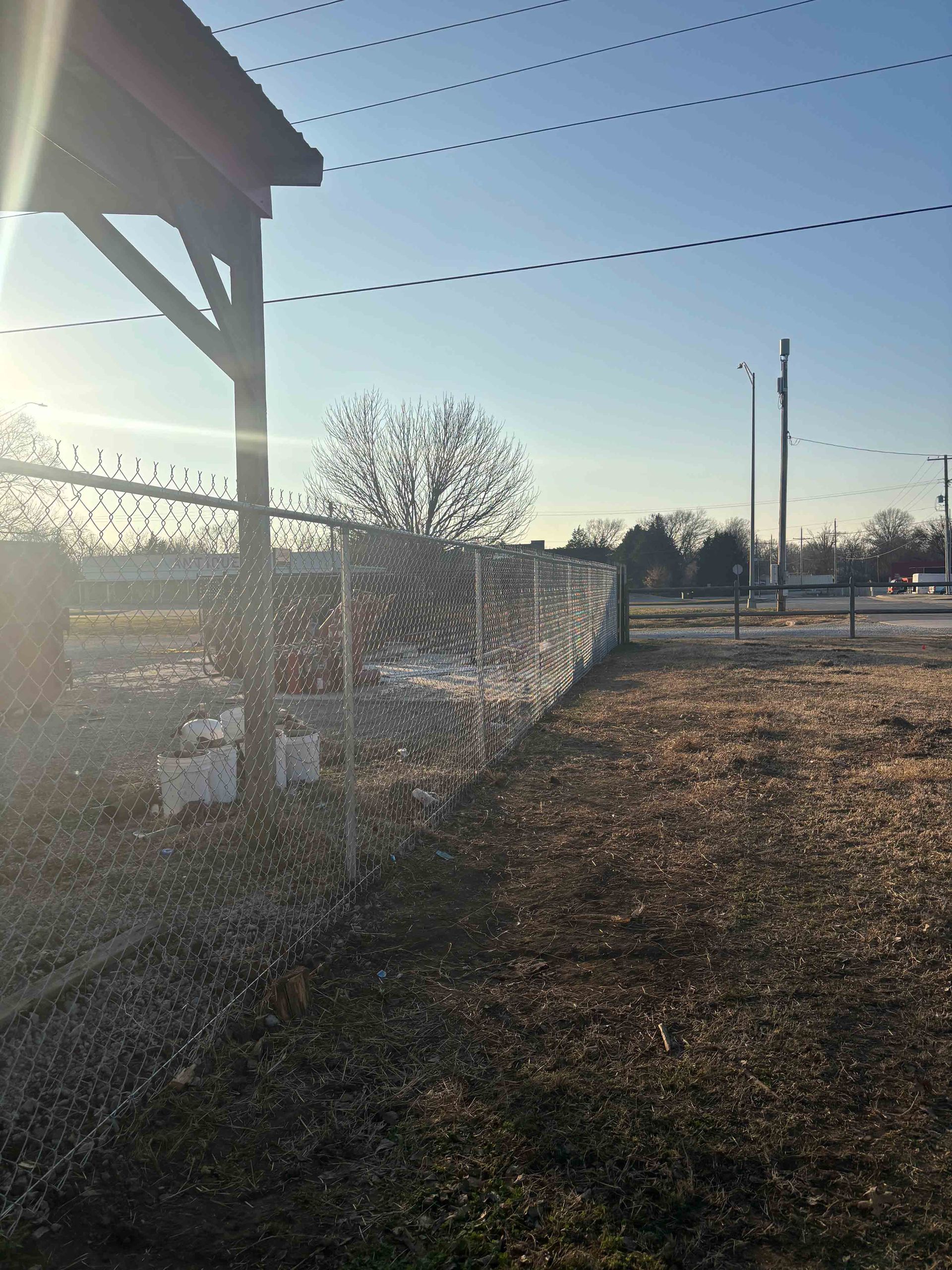 Sunlit chain-link fence beside a barn and leafless trees in a rural field
