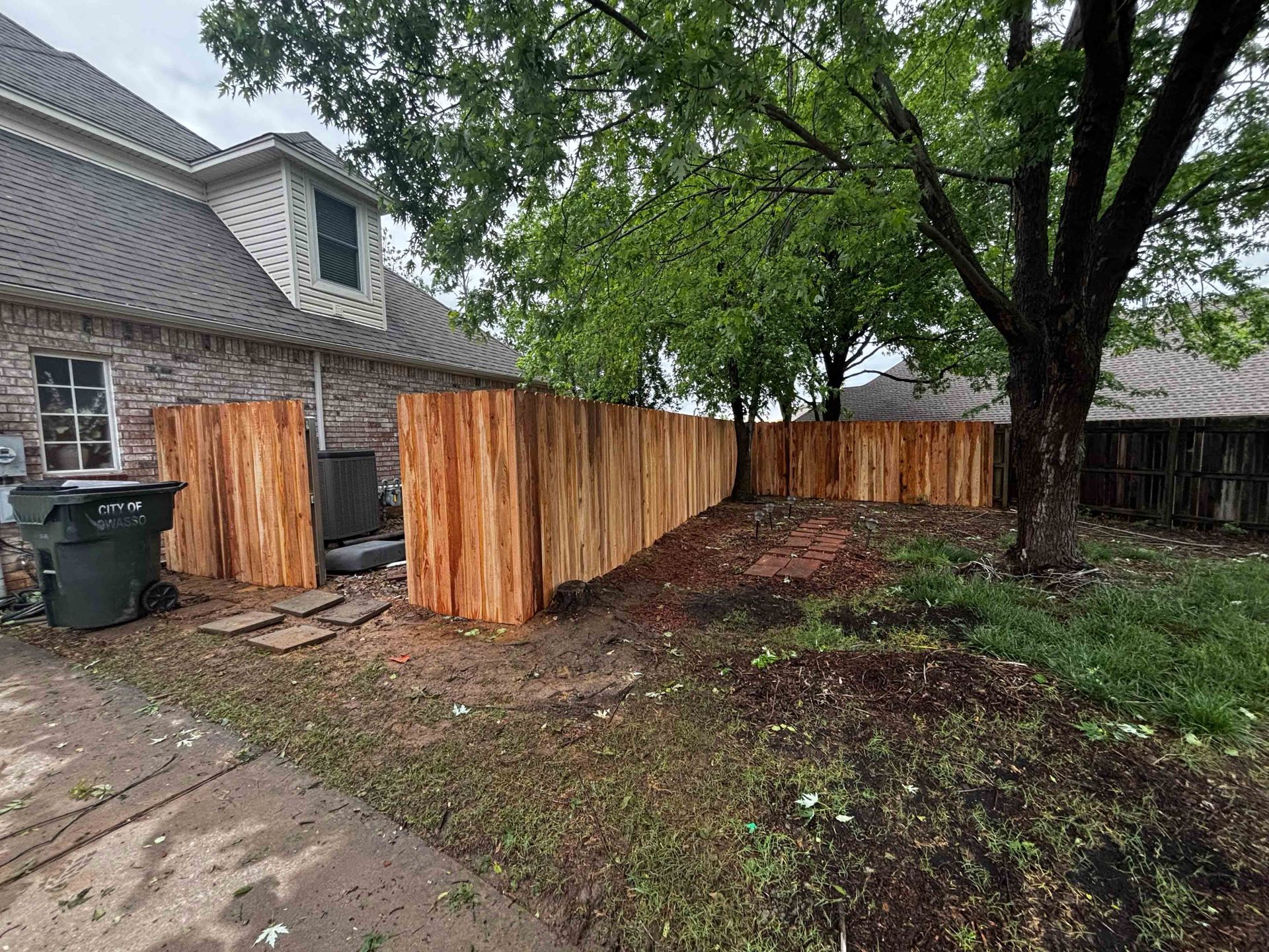 Wooden privacy fence beside a brick house, with a trash bin and a wet driveway in front.