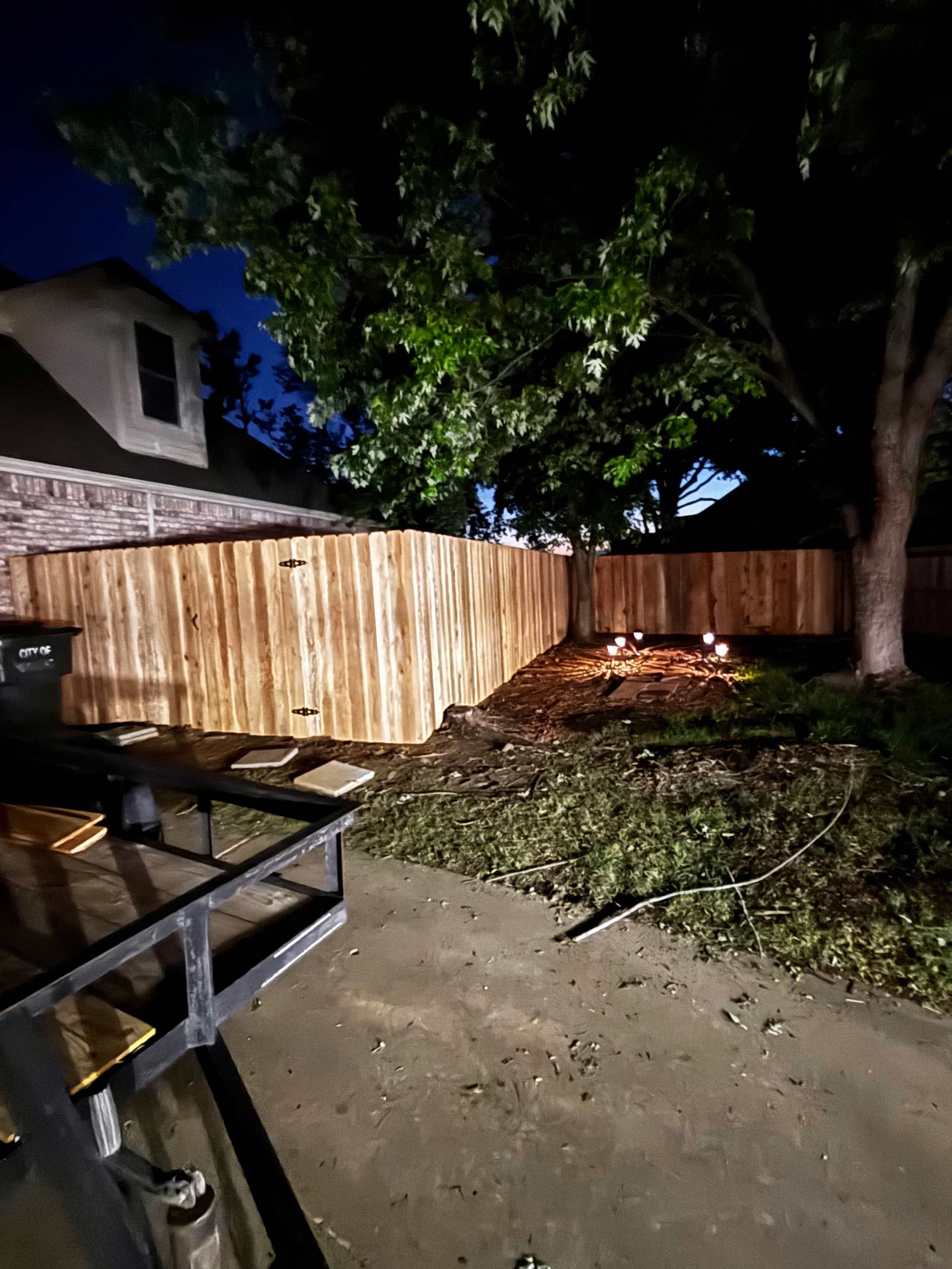 Backyard at night with a new wooden fence, trees, and a patio table in the foreground.