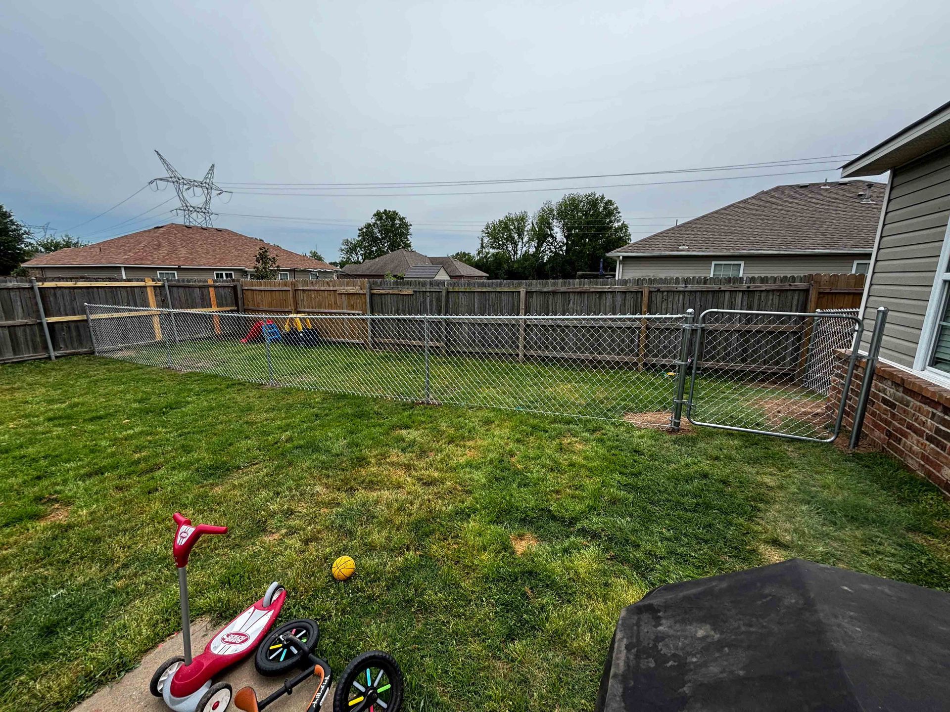 Backyard with grass, a tricycle and scooter, fenced yard, and neighboring houses under cloudy sky